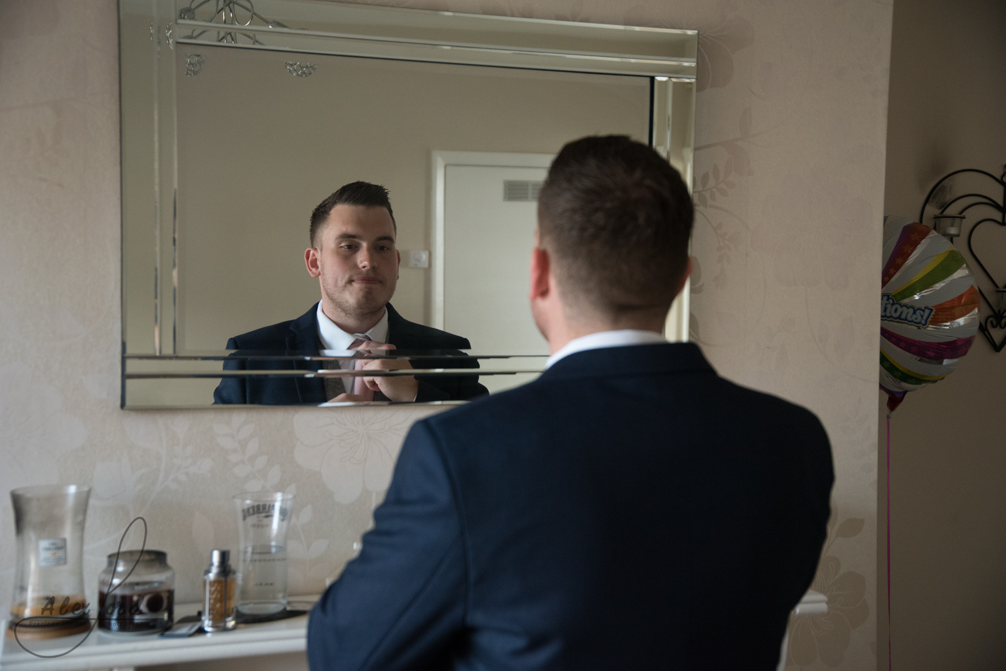 The groom dressed in a blue suit check his tie in the mirror before he leaves for his wedding ceremony