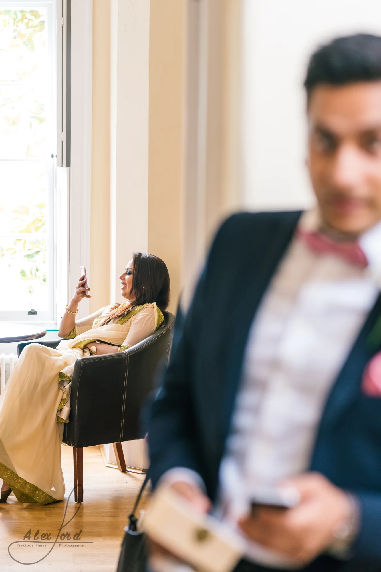 Last wedding guest dressed in gold sits on a chair in a sunlit room, partly hidden needing a man in a suit in the foreground