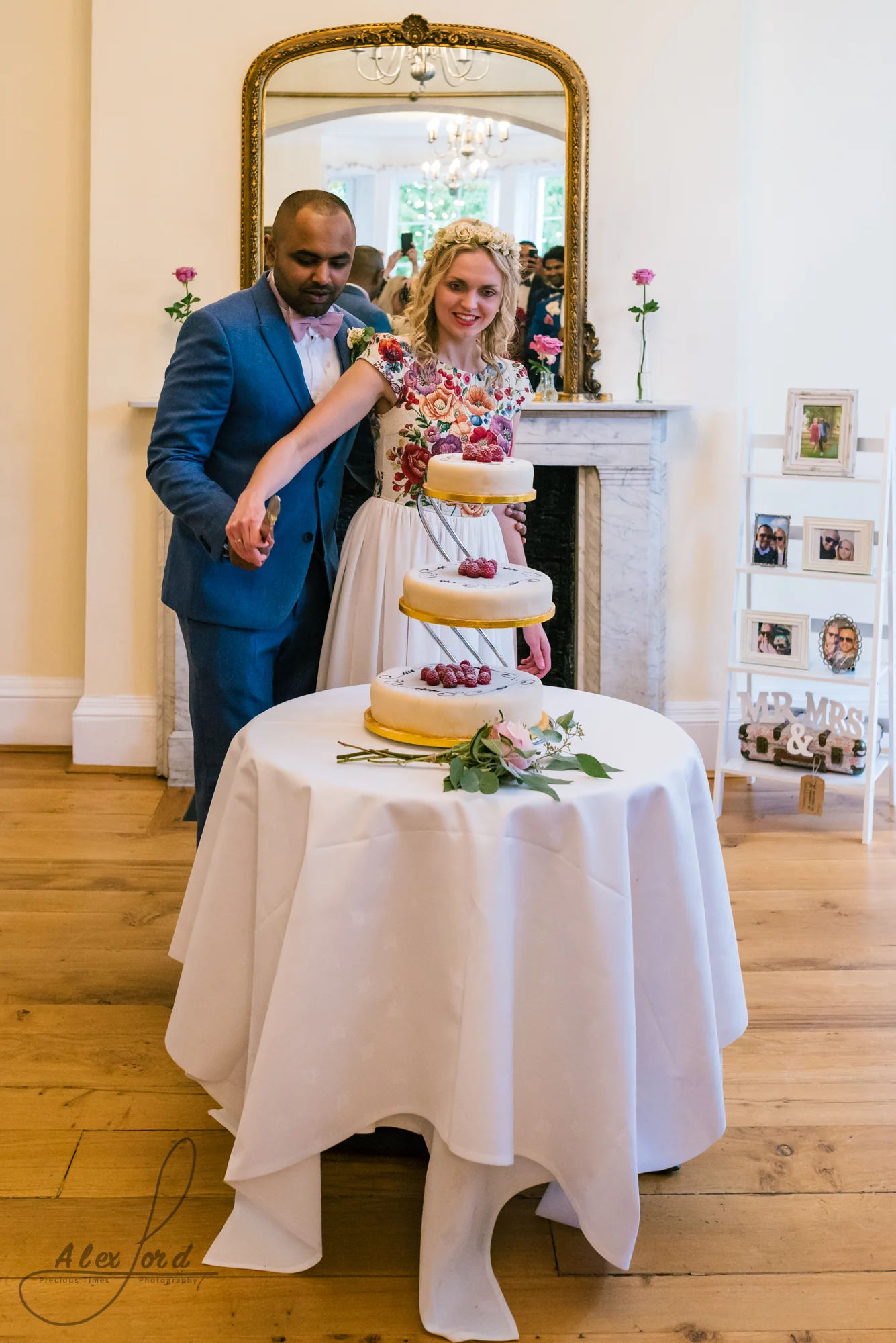 The bride and groom pose in front of a large mirror to cut their elegant wedding cake