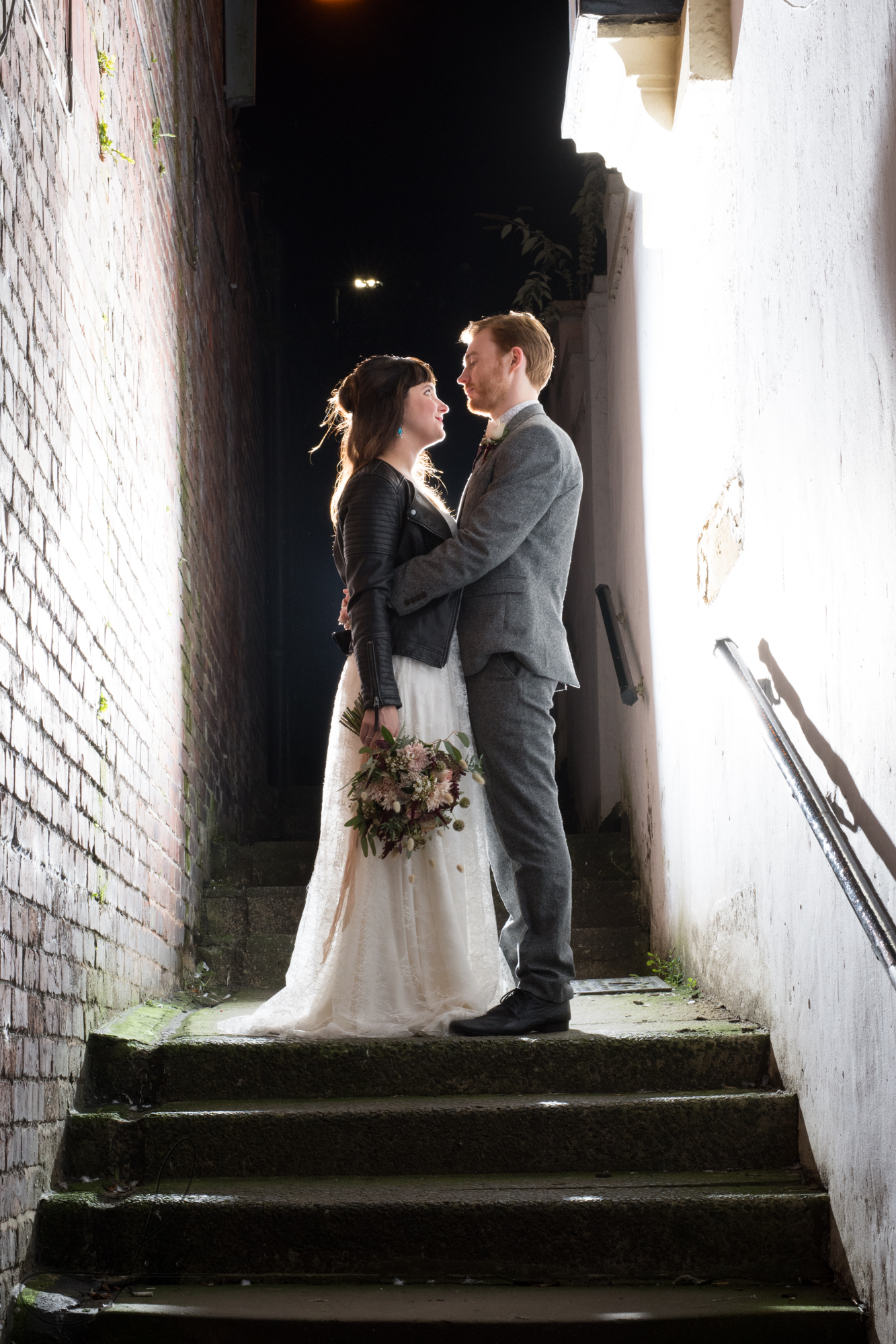 bride and groom stand on steps outside their venue for a photo with the flash behind them; they are looking at each other