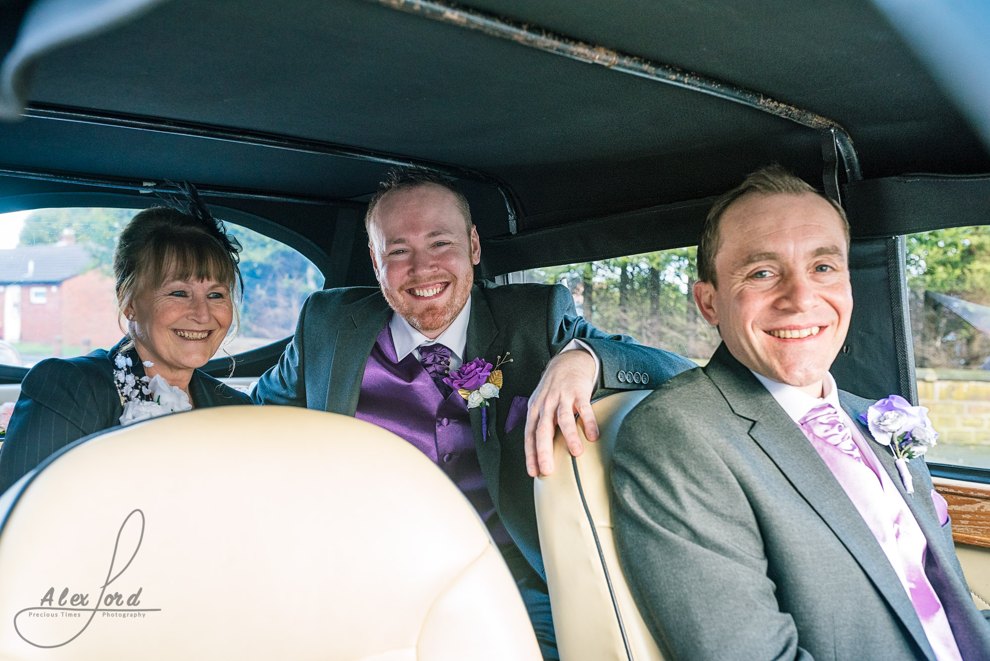 the groom sits in the wedding car with his mum and dad. The groom is in the back seat of the car