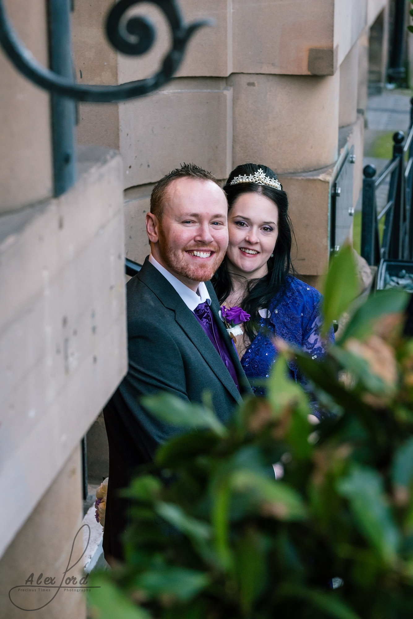 bride dressed in a purple dress and the groom in a dark grey suit stand outside their wedding venue