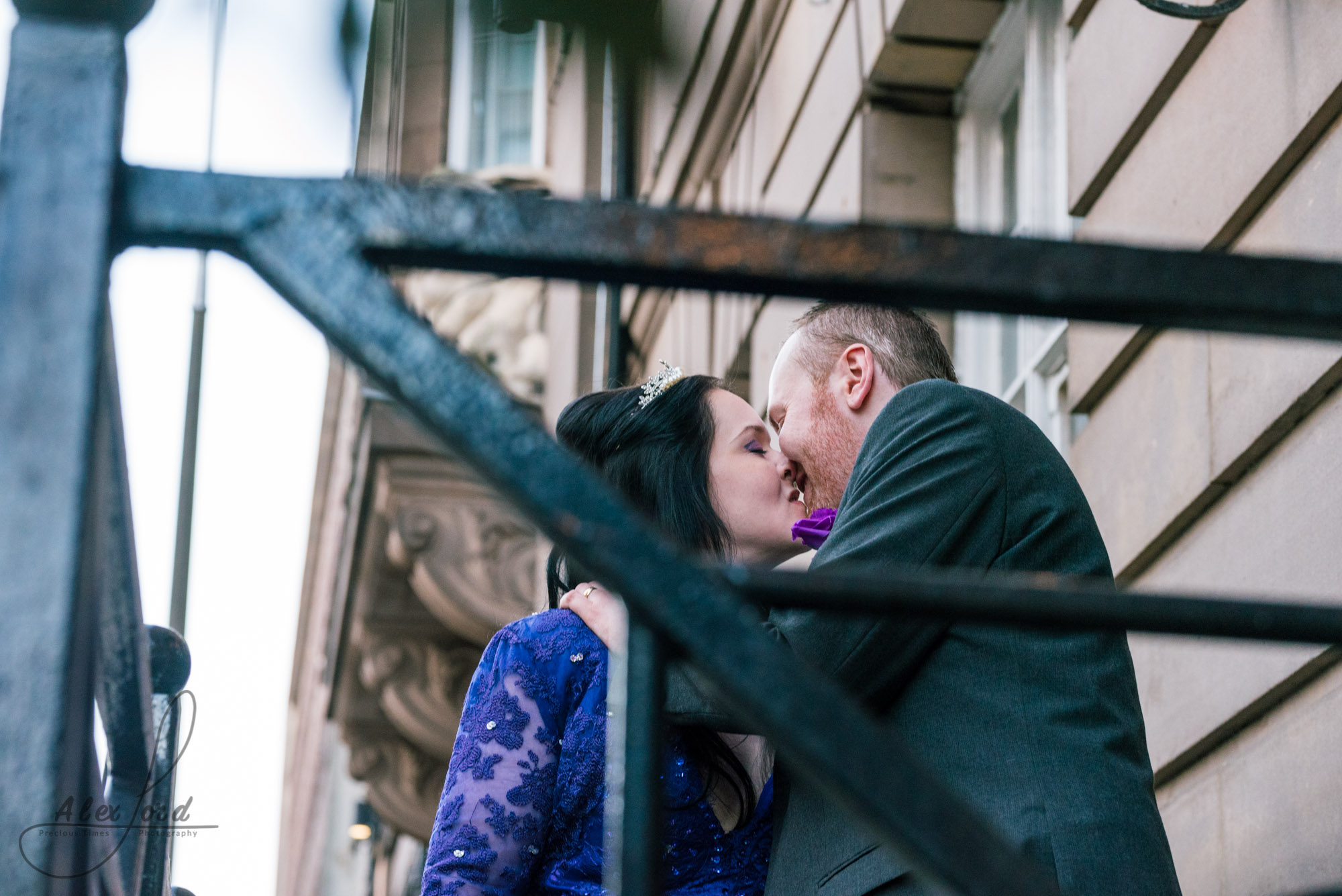 bride dressed in a purple dress and the groom in a dark grey suit stand outside their wedding venue