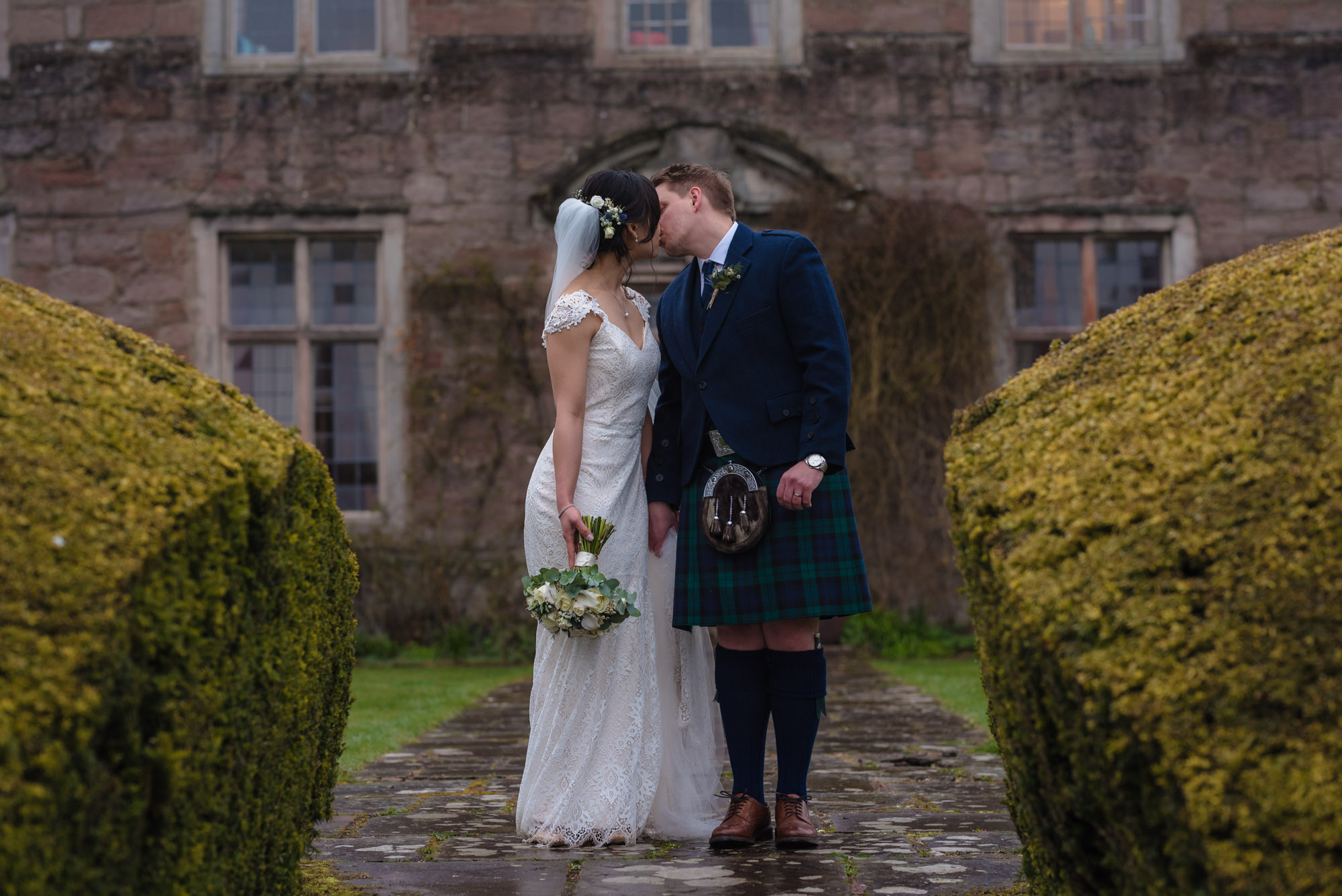 Bride and groom stand for a kiss in front of a damp Askham Hall.