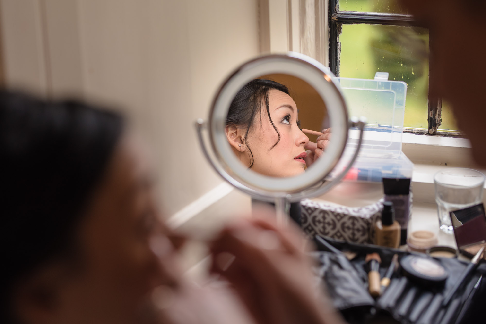 Bride having her make up done viewed through a round mirror in the window.