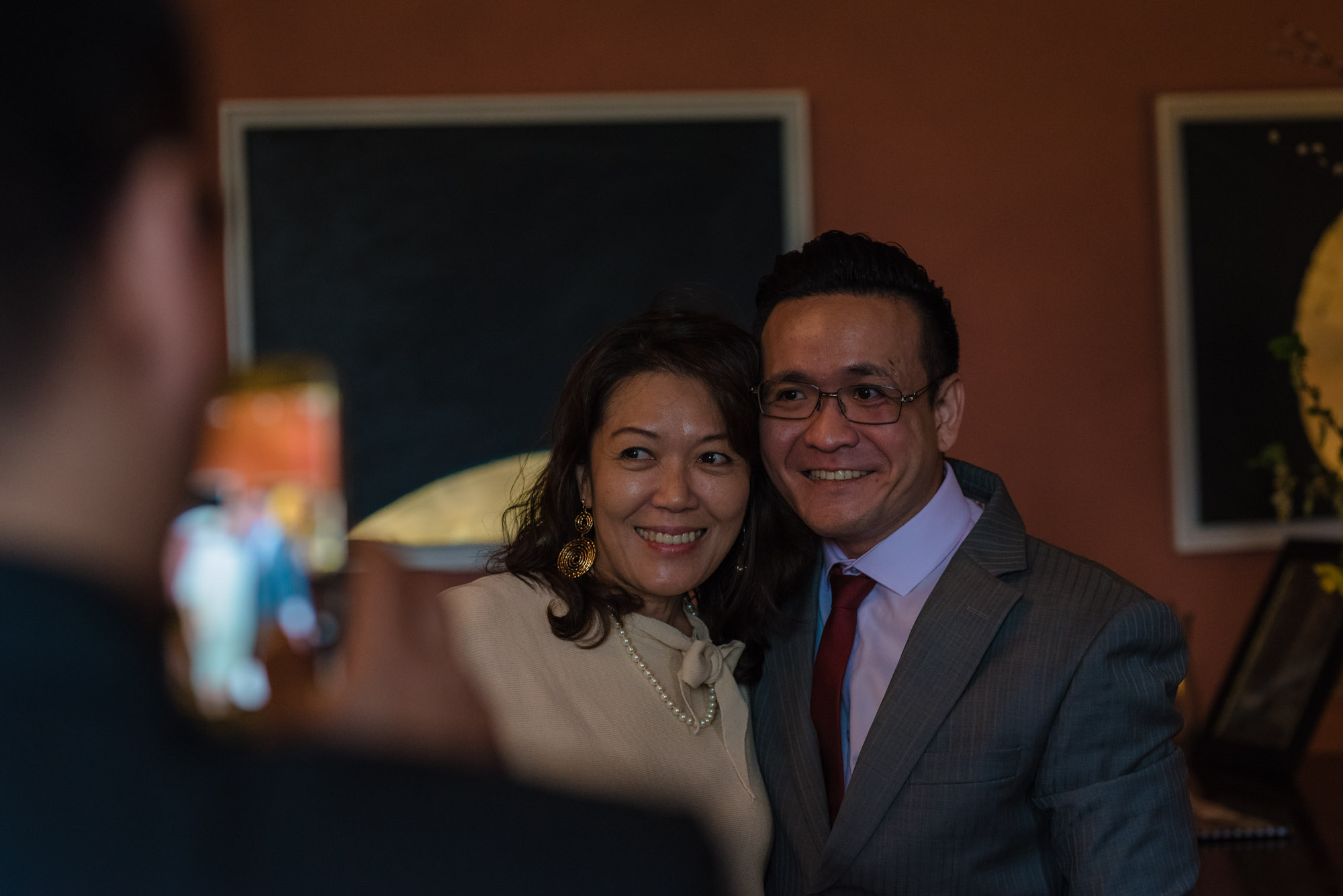 Guests stand against he wall while one of their friends takes a photo of them on their phone.