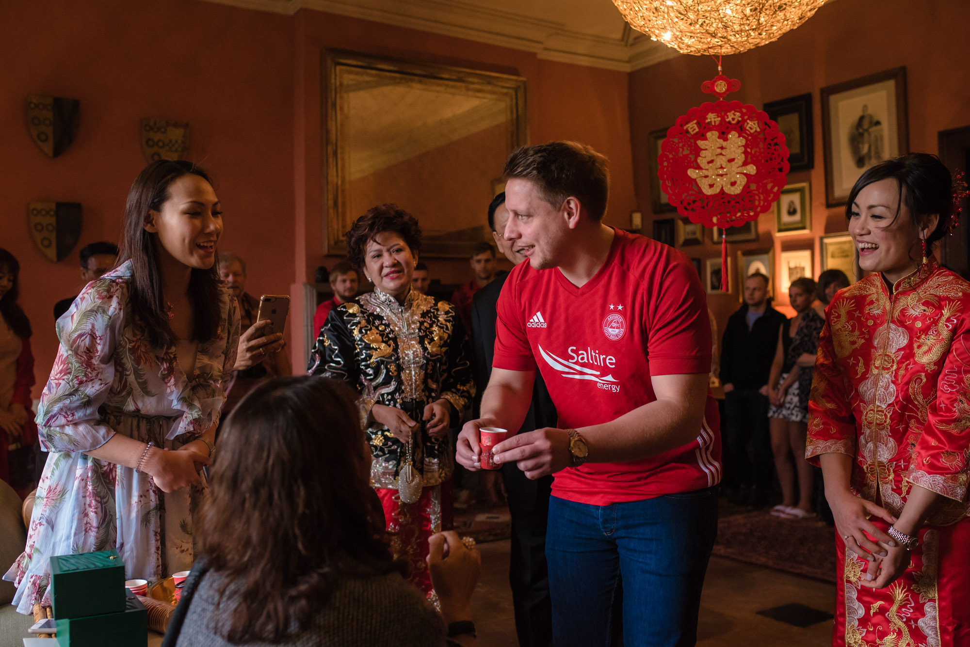 The groom in a red top presents tea to a seated member of the wedding party.