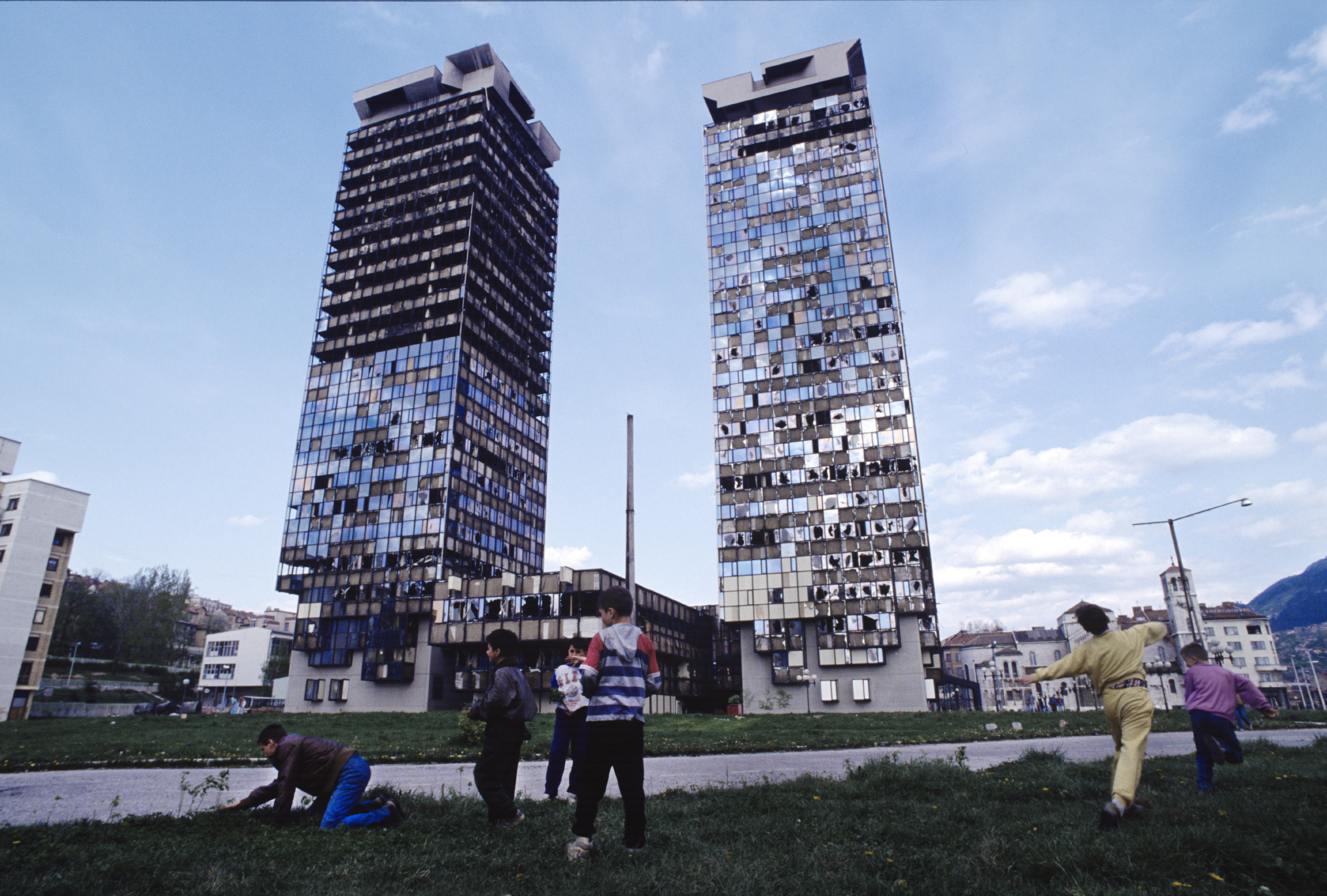  A man collects flowers and wild grass to eat while children play in front of the "Momo" and "Uzeir" towers on Sniper Alley in downtown Sarajevo. 1995 