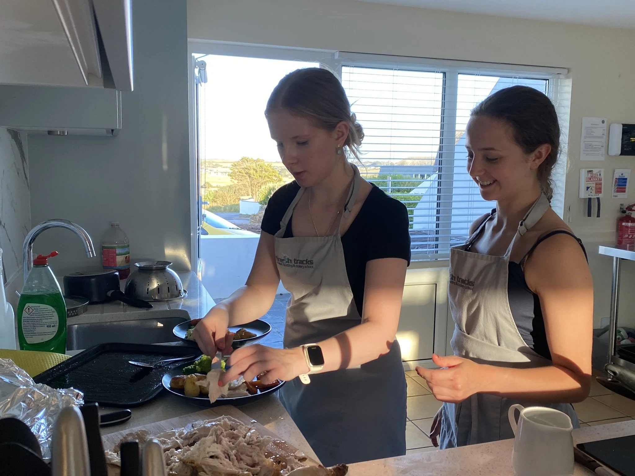 Two women are preparing food in a modern kitchen, wearing aprons and smiling. One woman is serving food onto a plate, while the other is watching with a smile. There are various kitchen items and ingredients on the counter, and large windows with blinds in the background.