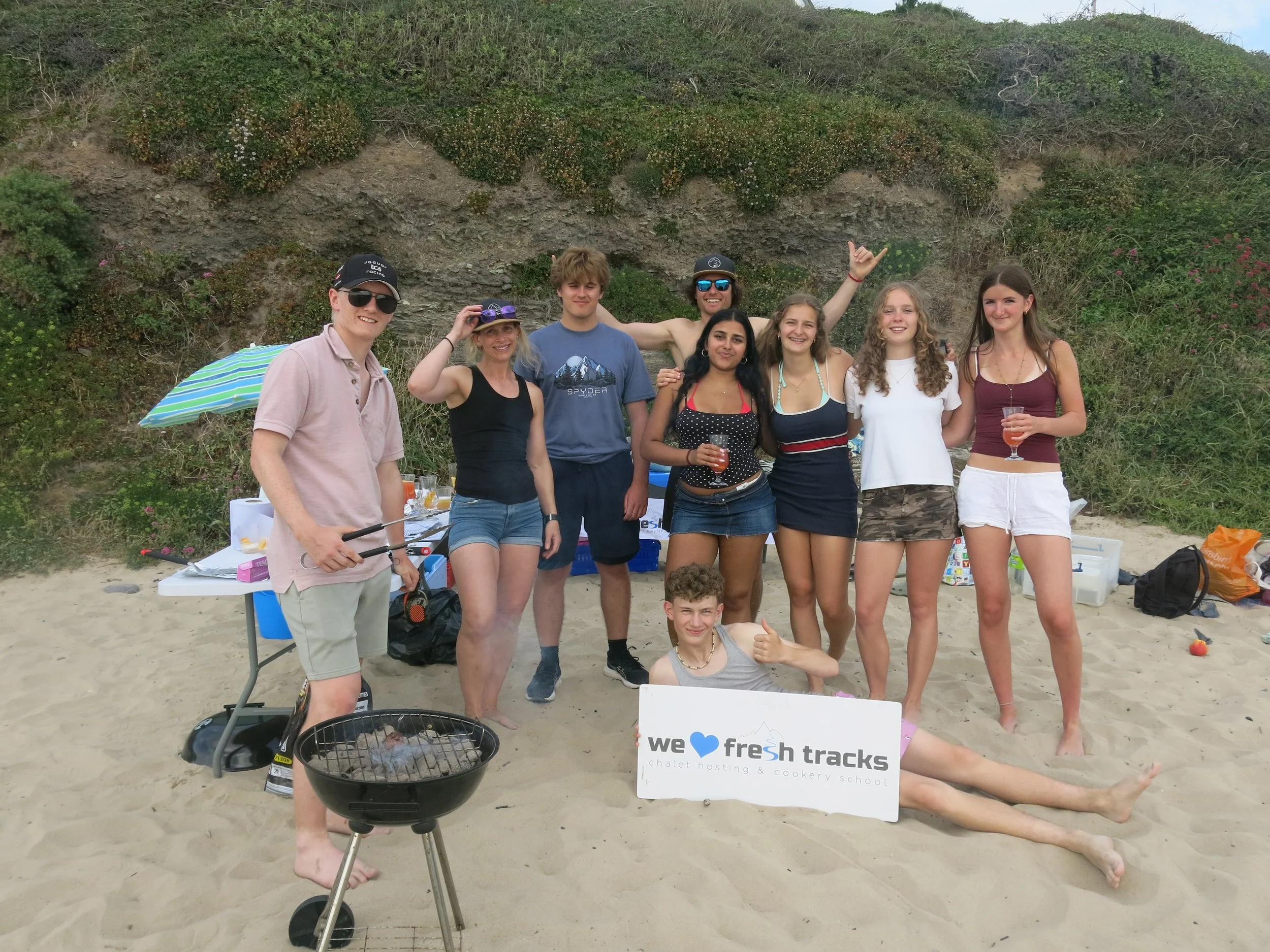Group of eight young adults standing on a beach, smiling, holding drinks, with a hillside with shrubs behind them. One person is grilling, and a sign in front reads 'we ♥️ fresh tracks, chalet hosting & cookery school'.