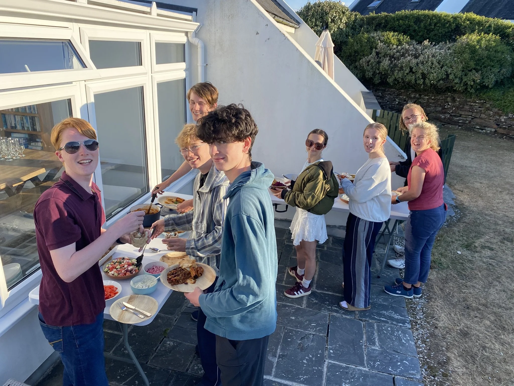 Young people enjoying an outdoor buffet on a balcony, serving themselves food with bowls and plates, with greenery and a white building in the background on a sunny day.