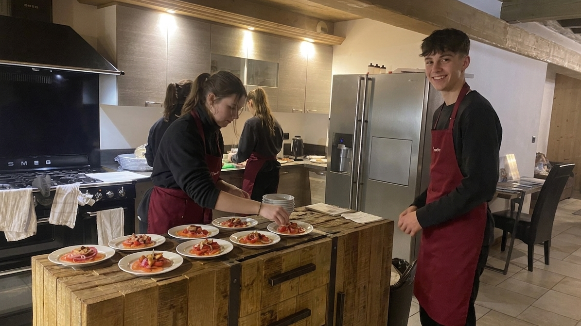 Young guy chalet hosting in the kitchen helping a chalet girl prepare and plate up the evening meal in a warm and cozy chalet kitchen.