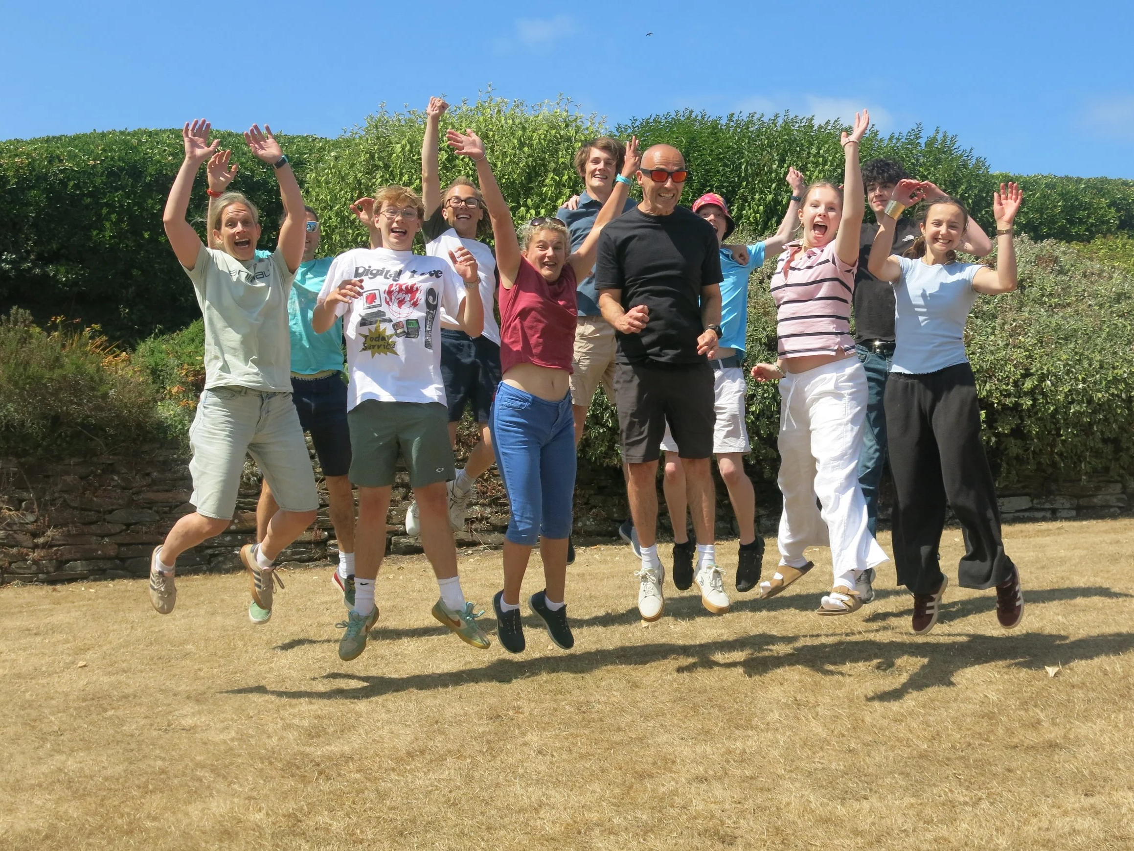 Group of people jumping and smiling outdoors on a sunny day with green bushes and a blue sky in the background.