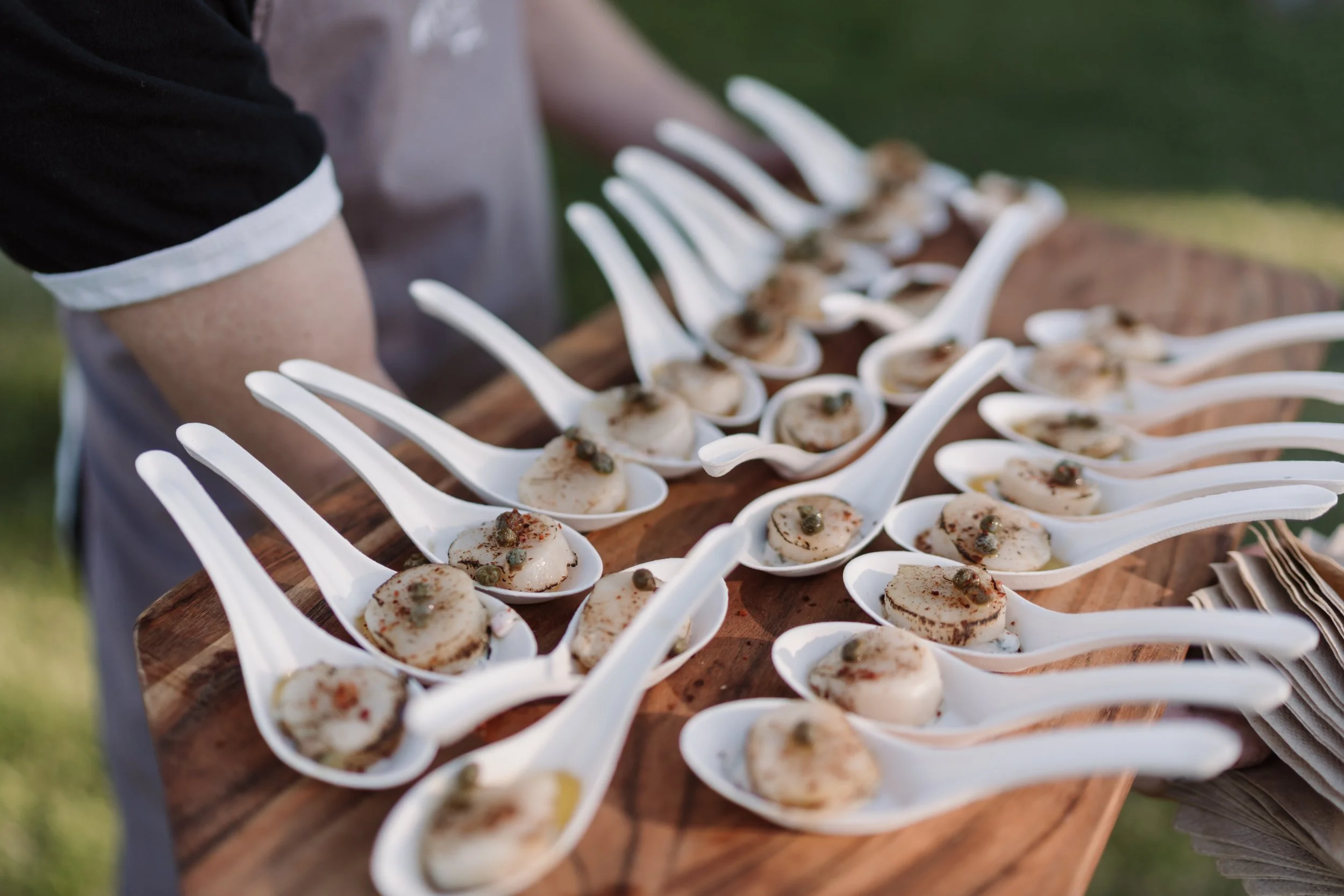 A person serving small bites on white ceramic spoons arranged on a wooden tray outdoors.