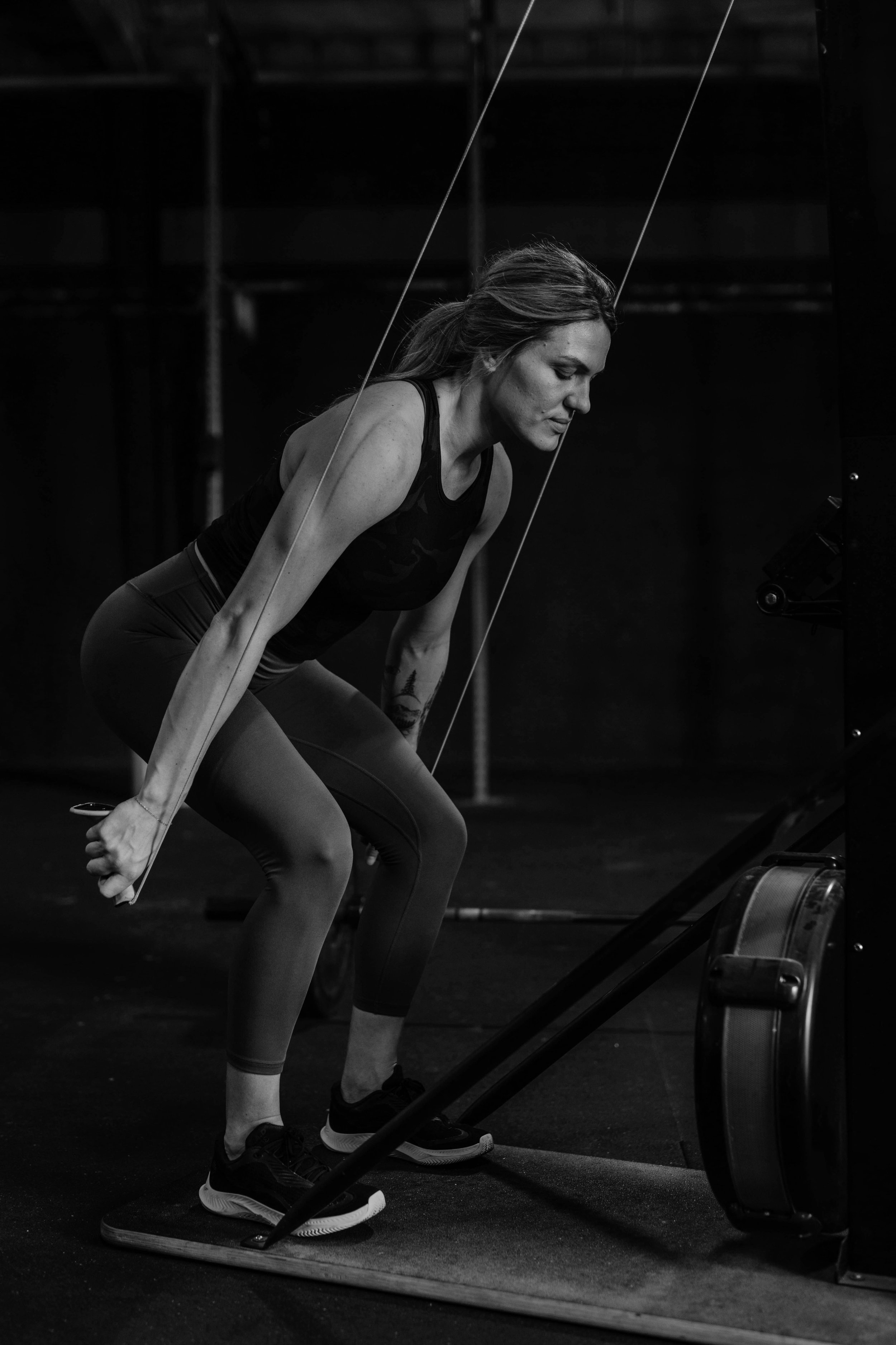 A woman with athletic build wearing a black tank top and leggings, demonstrating a rowing exercise on a rowing machine in a gym with dark background.