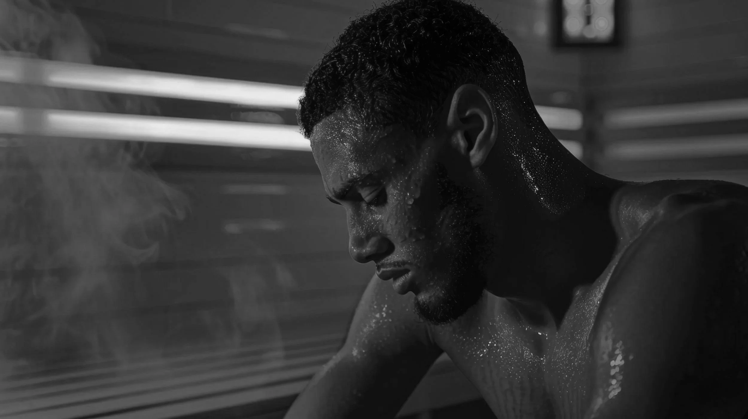 A man with water droplets on his face and upper body, looking down with a serious expression in a room with horizontal lighting.