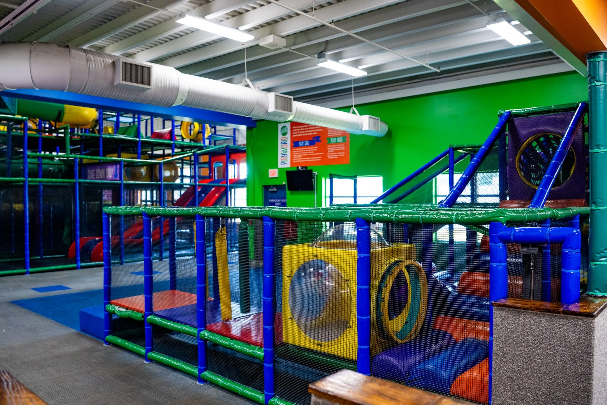 Colorful indoor children's play area with slides, tunnels, and climbing structures, enclosed by mesh fencing.