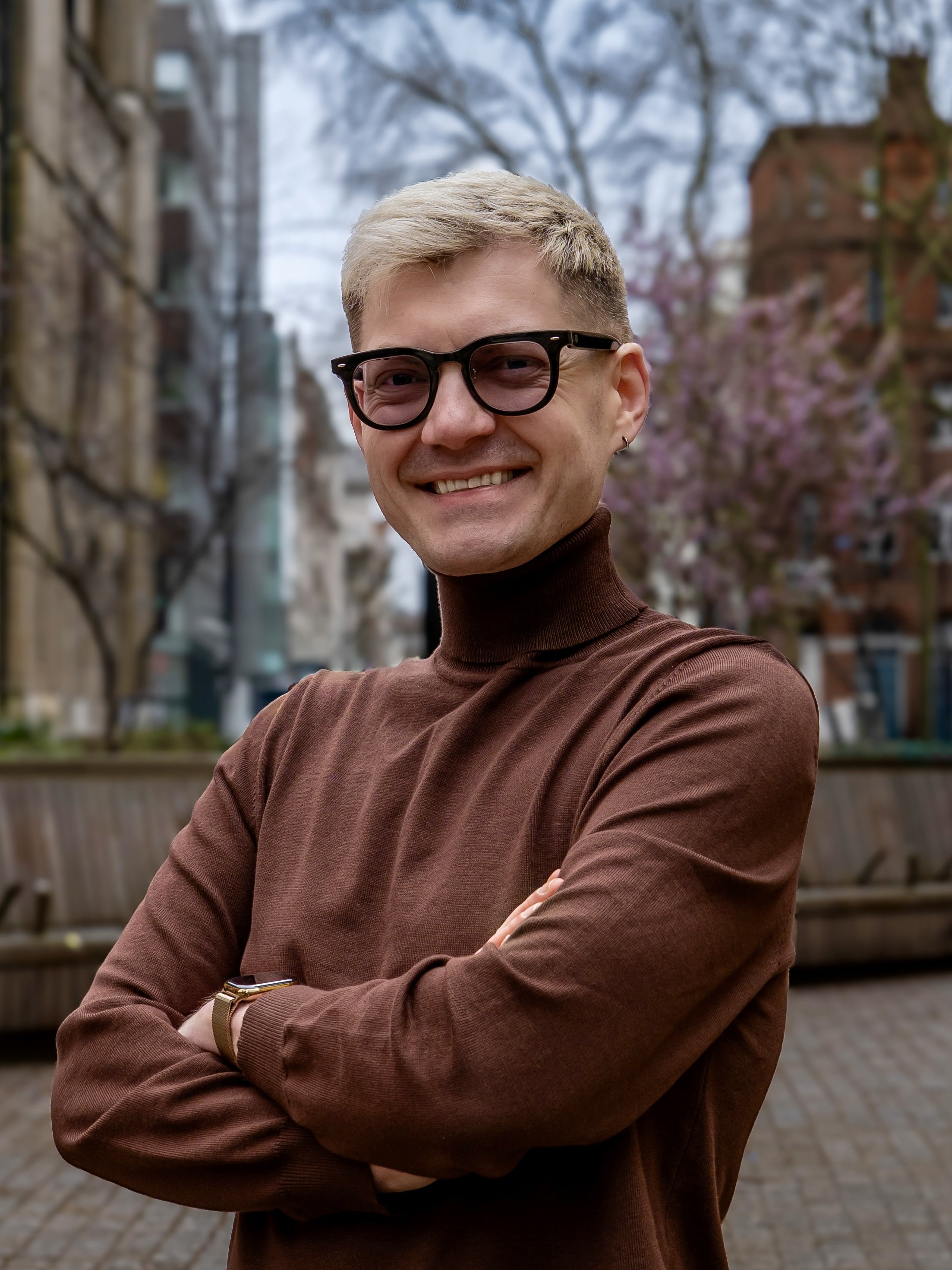 A young man with blonde hair, wearing glasses and a brown turtleneck, smiling with arms crossed outdoors with bare trees and buildings in the background.