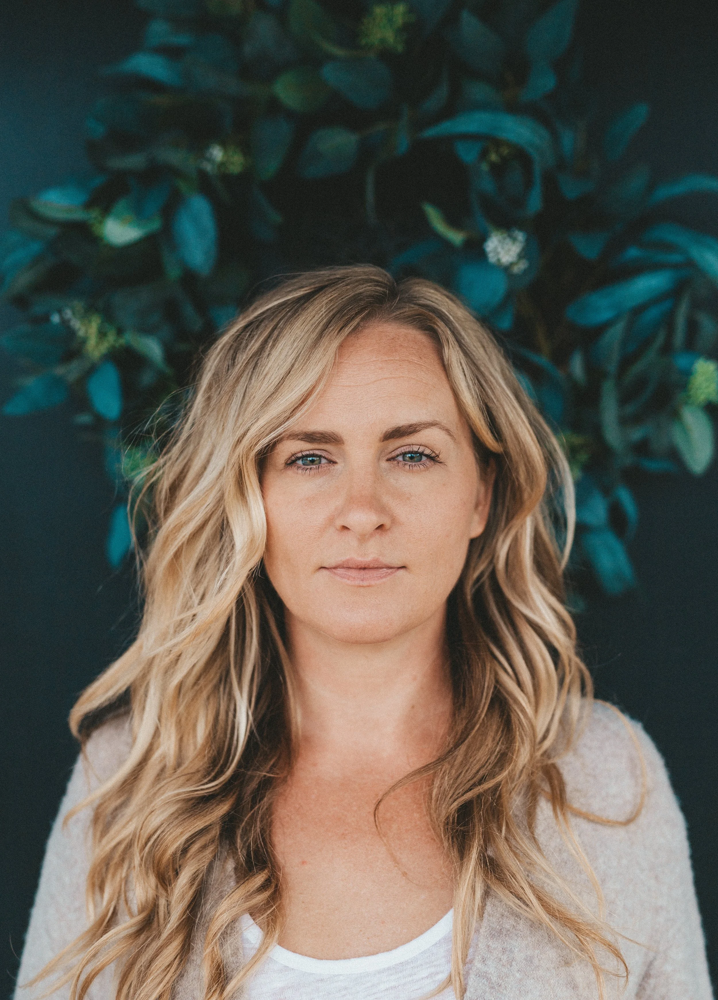 Woman with long blonde hair standing in front of green leafy background.