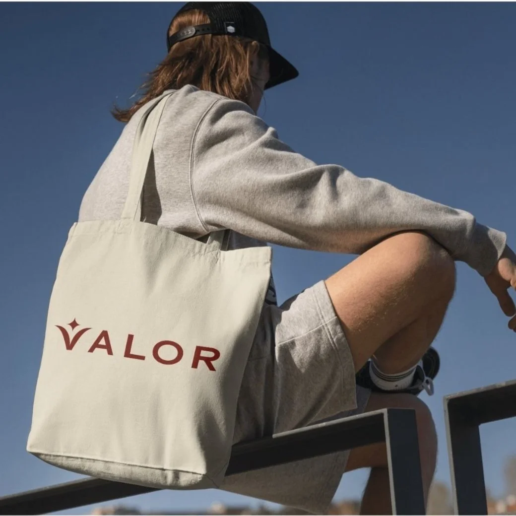Person wearing gray athletic attire and black cap, sitting outdoors with a tote bag labeled 'VALOR.'