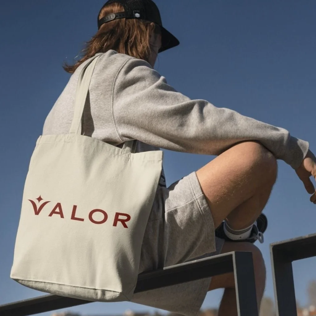 Person wearing a cap and gray sweatshirt, sitting with a canvas tote bag labeled "VALOR."