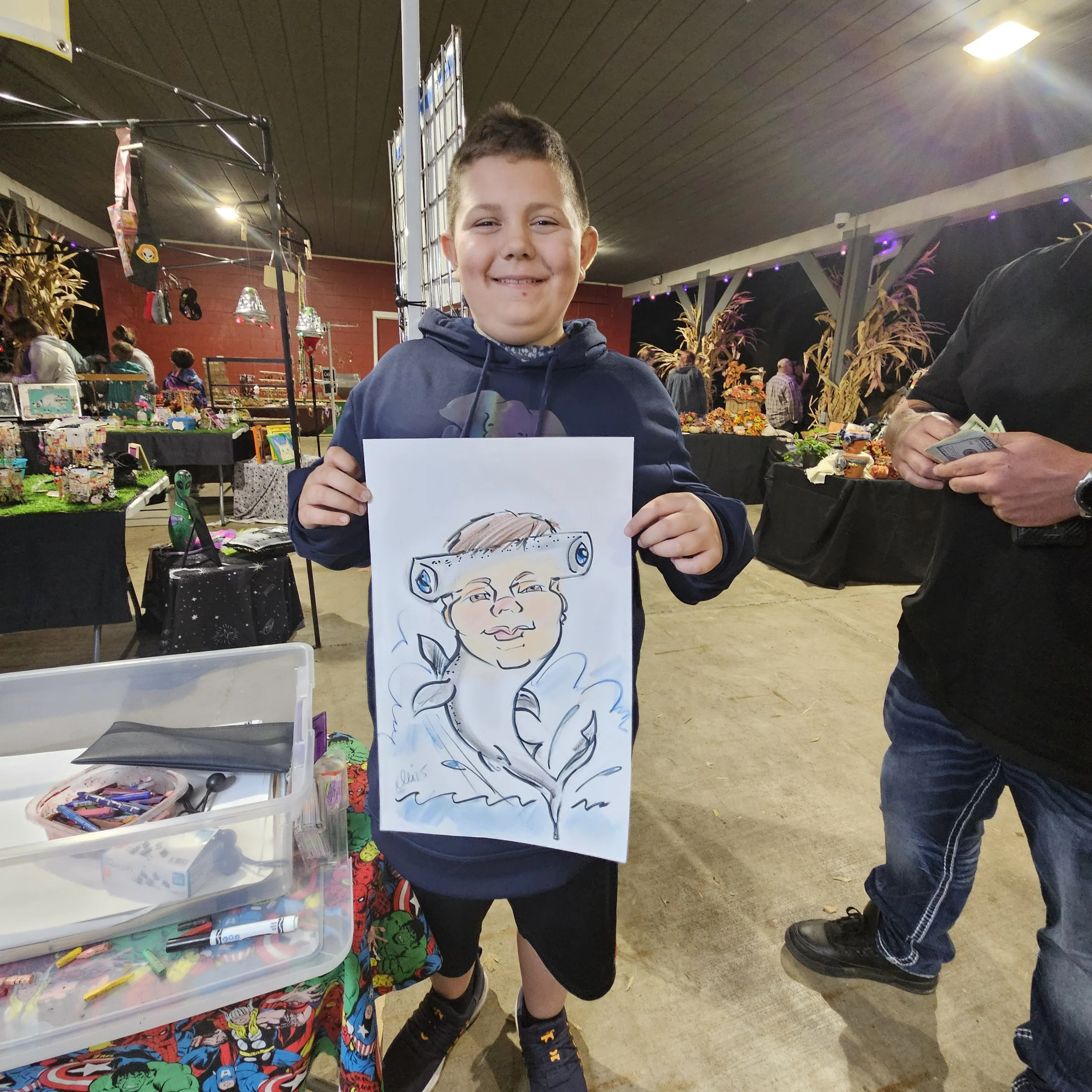 Young boy at an indoor craft fair holding a caricature drawing of himself wearing a hair roller.