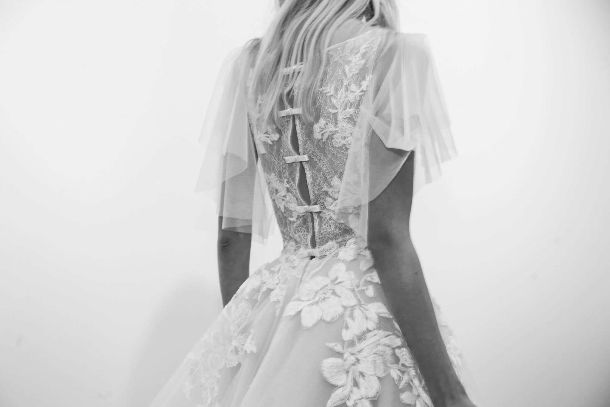 Bliss Monique Lhuilier shows a feminine wedding dress look with tailored bows and a sheer petal sleeve. 