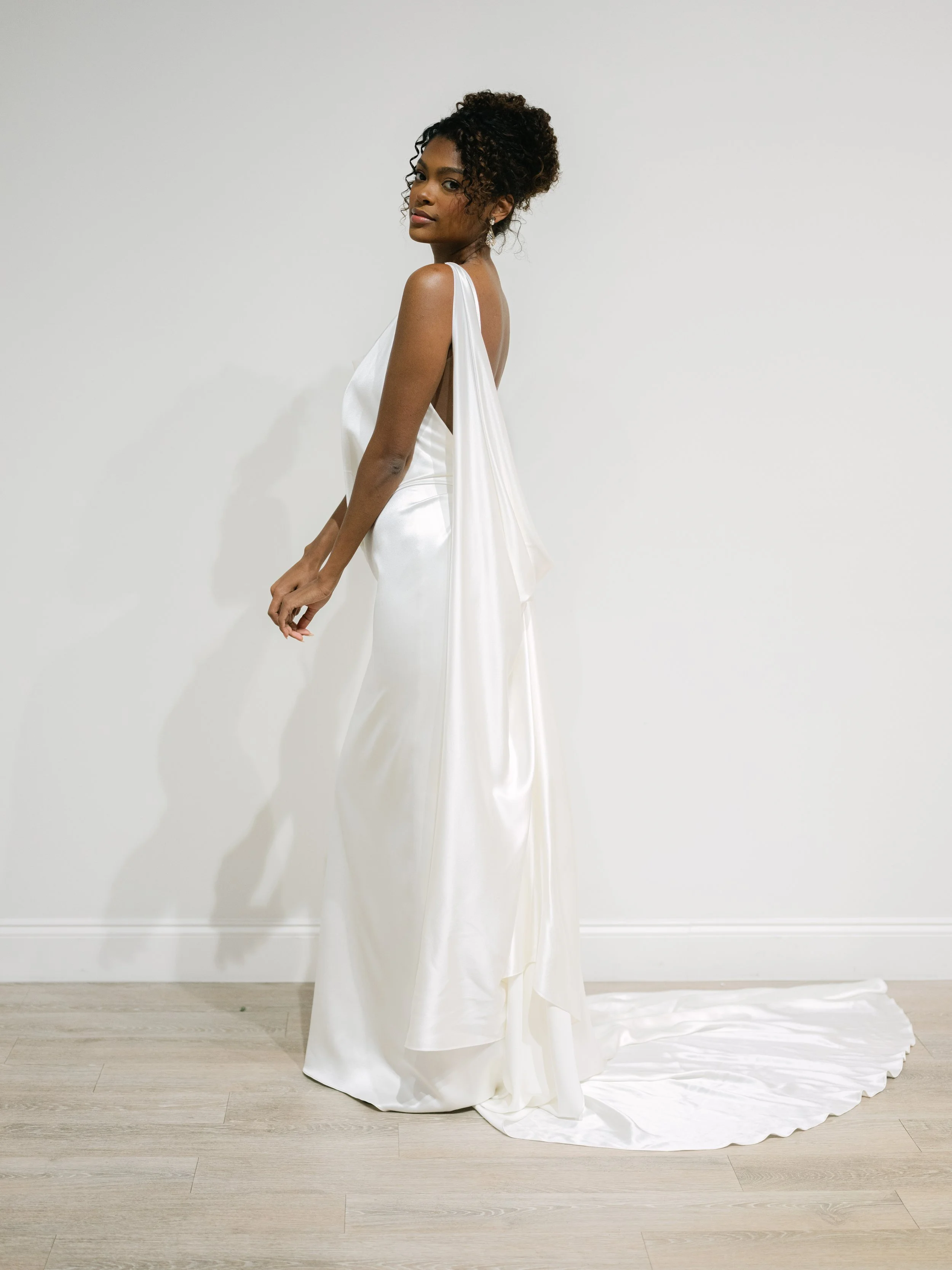 satin open back wedding dress with a flowing detachable cape. Plunging neckline wedding dress with open back. Satin wedding dress with open back and draping details