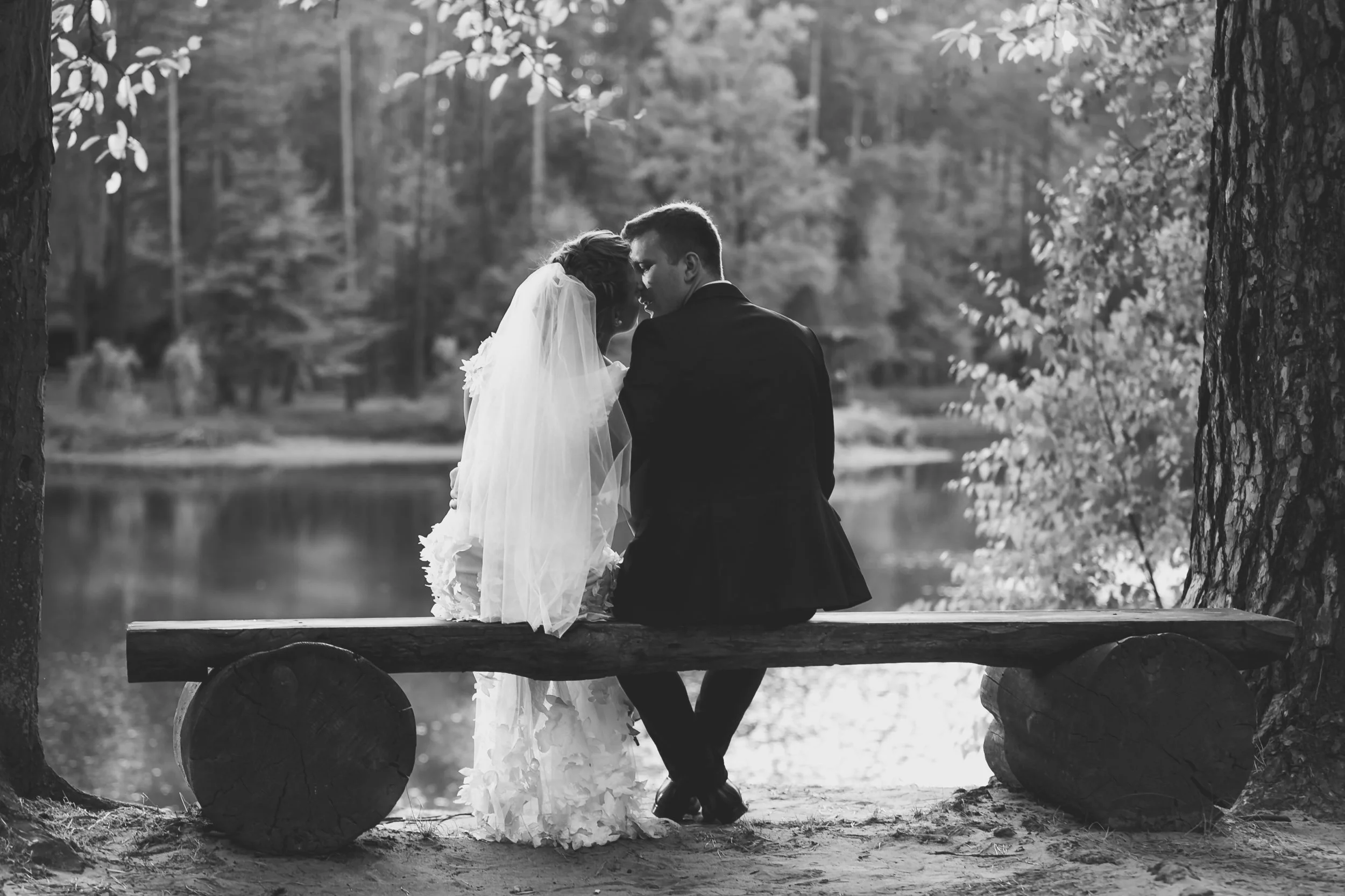 Monochrome photo of bride and groom kissing on bench at riverban..