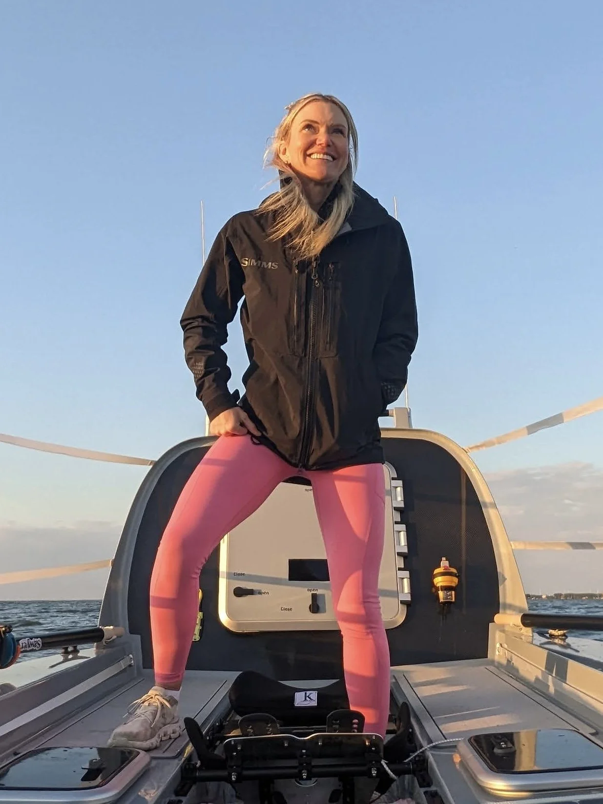 Woman standing on a boat, smiling, wearing a black jacket and pink leggings, with the sky and water in the background during sunset.