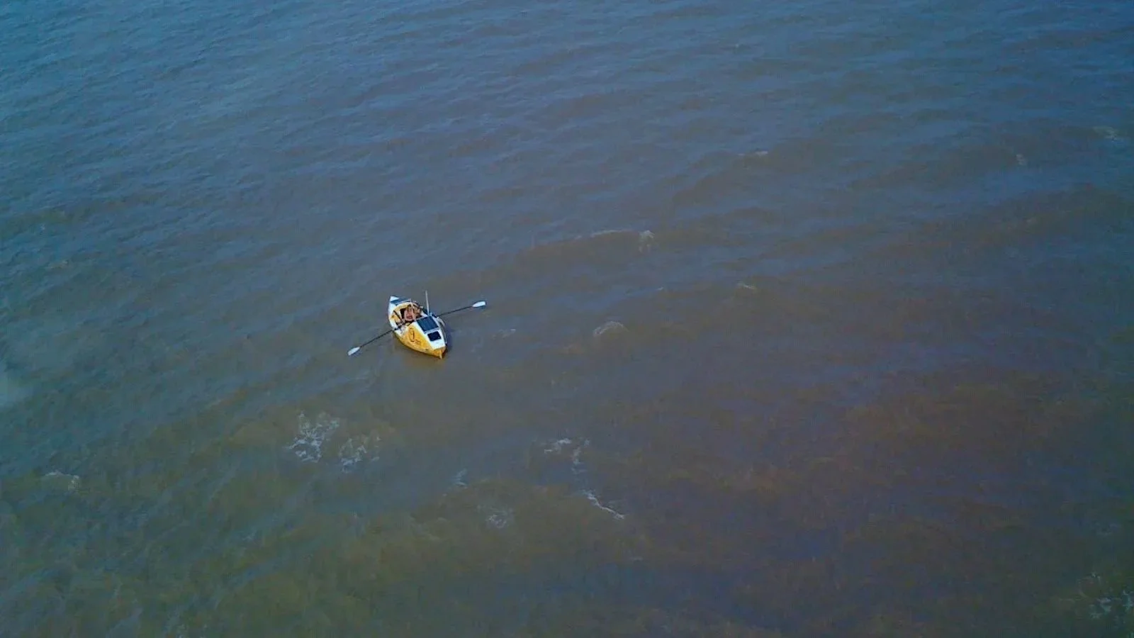 An aerial view of a small yellow and white boat with two passengers and two paddles on a calm, brownish body of water.