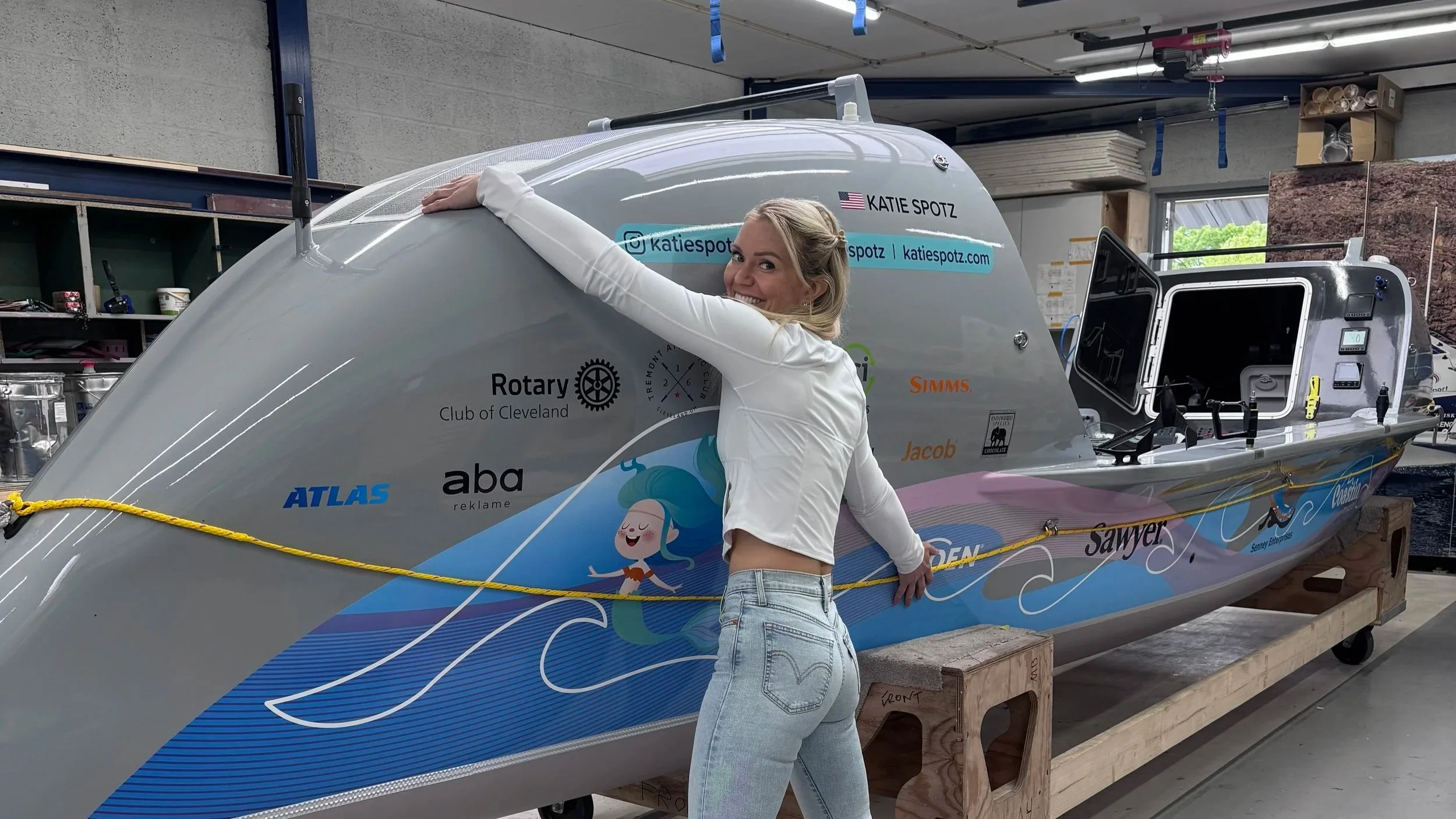 A woman in a white jacket and jeans is hugging a large, colorful boat inside a workshop or garage. The boat has various sponsor logos and a cartoon mermaid illustration on its side. The woman is smiling and looking at the camera.
