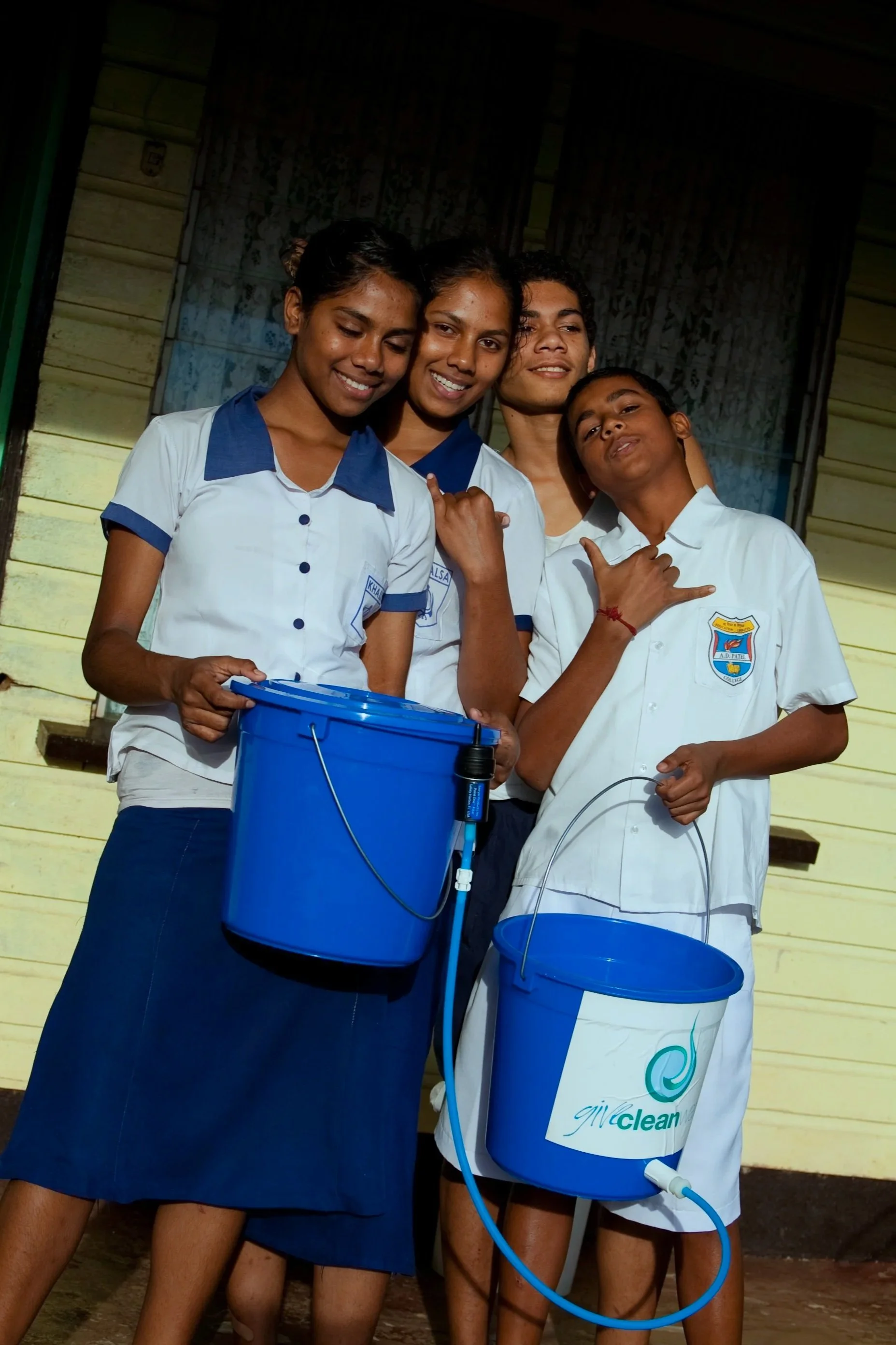 Group of four teenagers standing close together, smiling, holding blue buckets and a foam gun, standing outside a yellow wooden house.