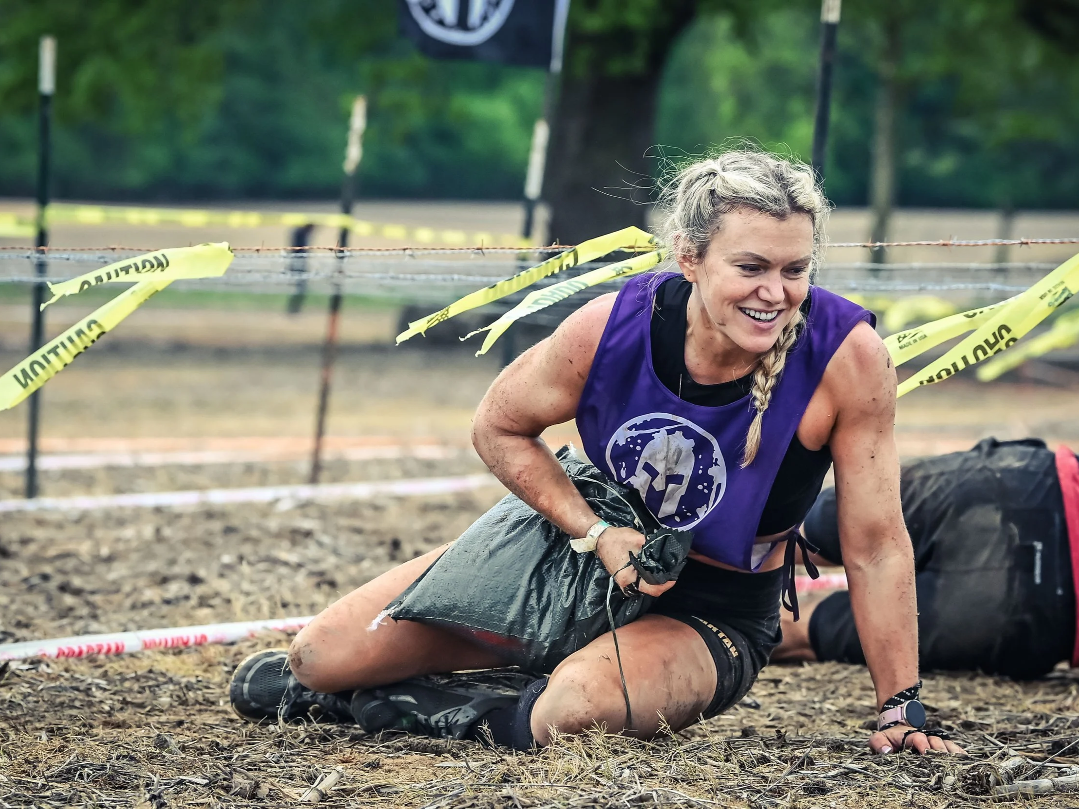 Female obstacle course competitor crawling on muddy ground during outdoor competition, surrounded by yellow caution tape and trees in the background.