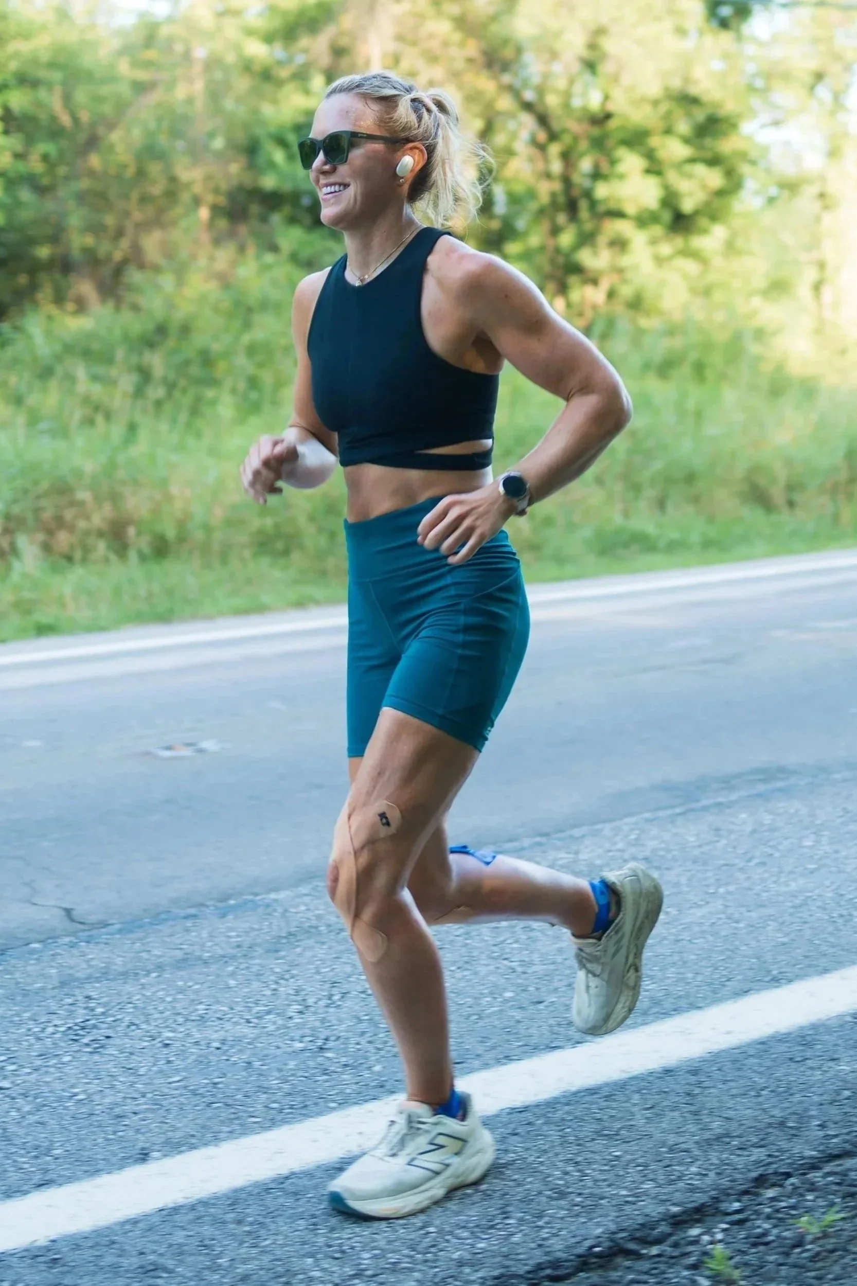 A woman running outdoors on a paved road with trees in the background. She has blonde hair tied back, wears sunglasses, a black sports bra, blue shorts, white running shoes, a watch, and wireless earbuds.