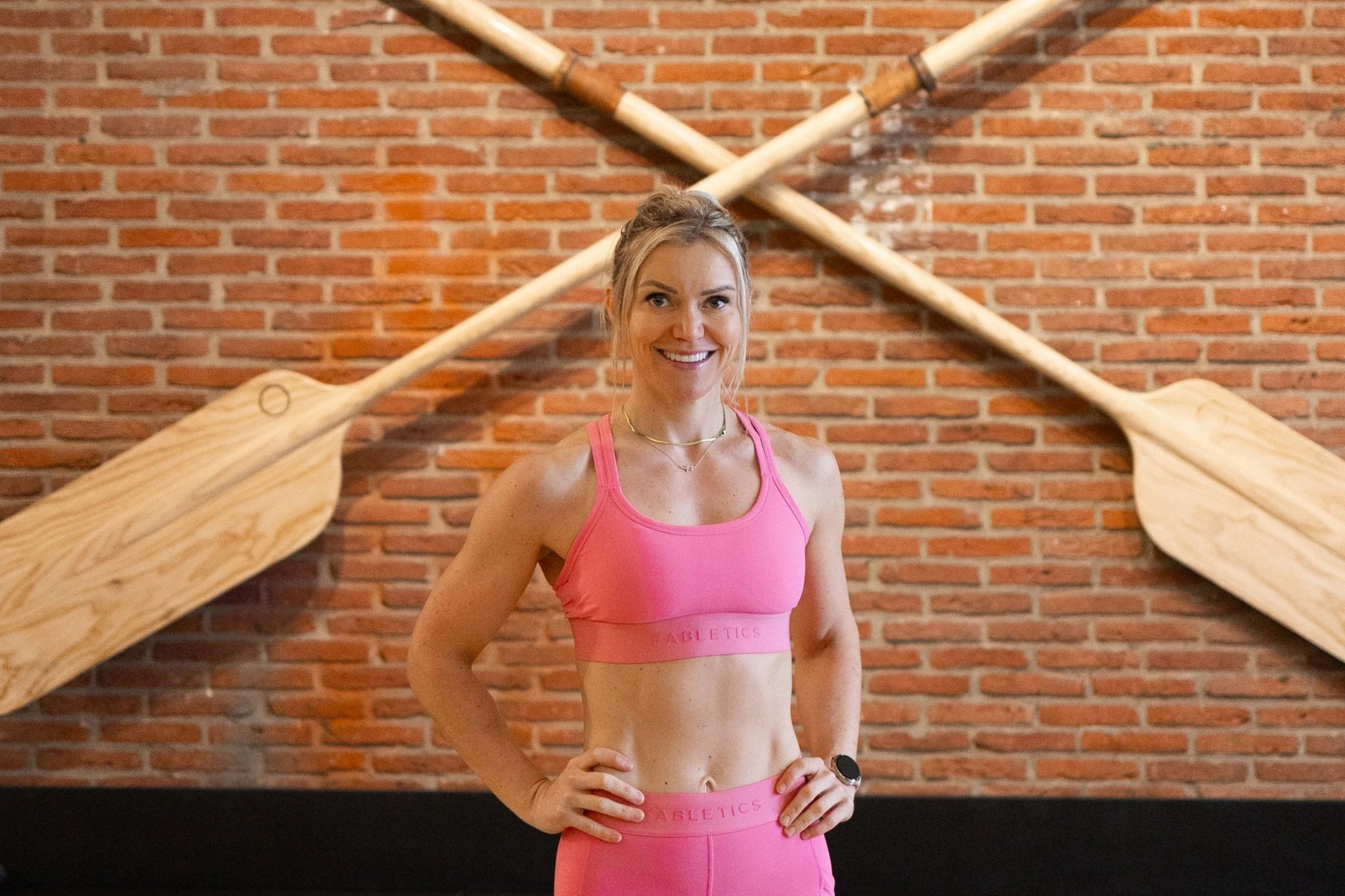 A woman in a pink sports bra and leggings standing confidently with her hand on her hip, smiling in front of a brick wall with crossed oars mounted behind her.
