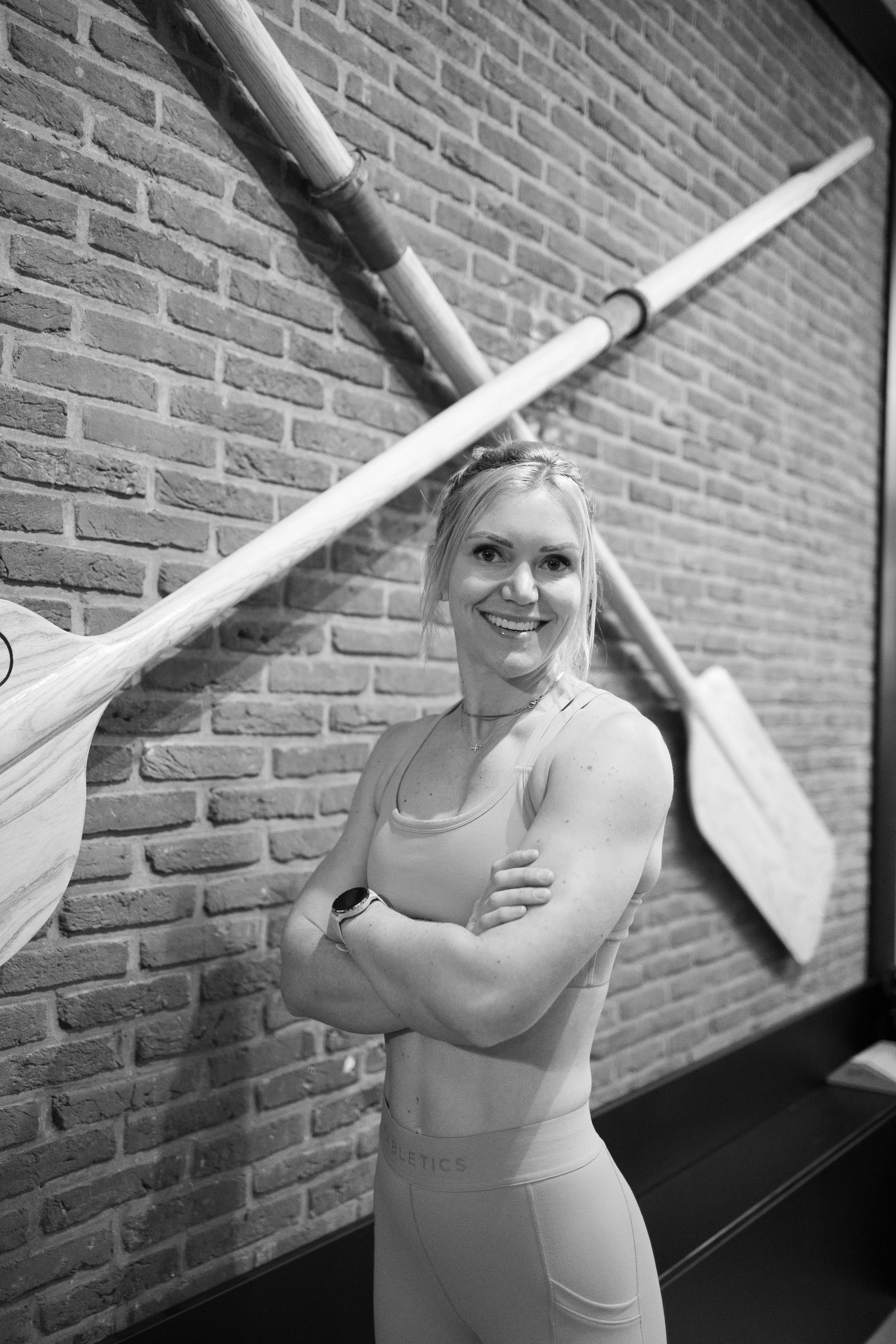 A smiling woman in athletic clothing posing with her arms crossed in front of a brick wall in a gym.