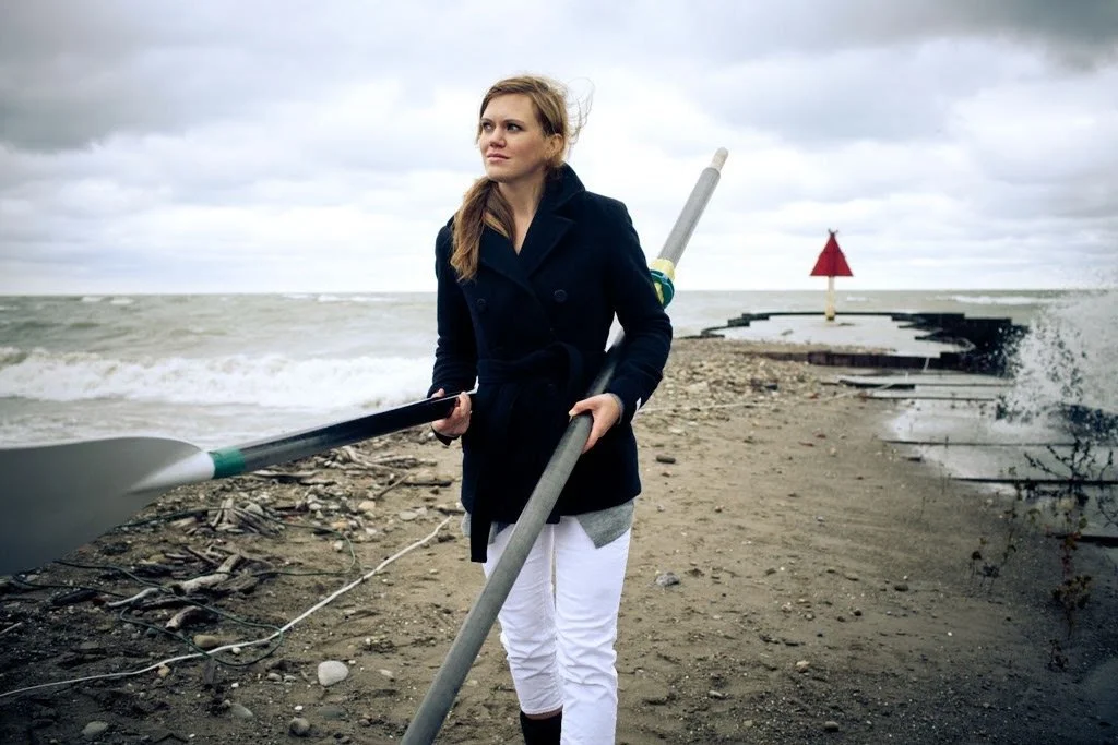 Woman in navy coat and white pants holding a paddle on a rocky beach with cloudy sky and a red marker in the background.