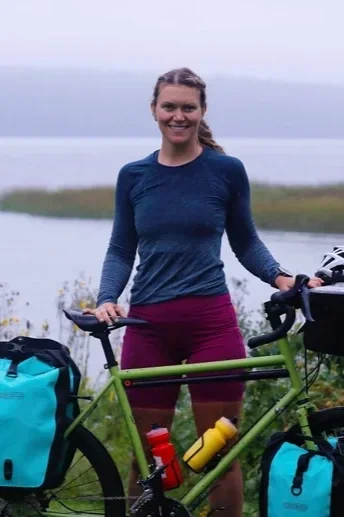 A woman standing outdoors with a green bicycle equipped with blue and yellow bags near a body of water during daytime.