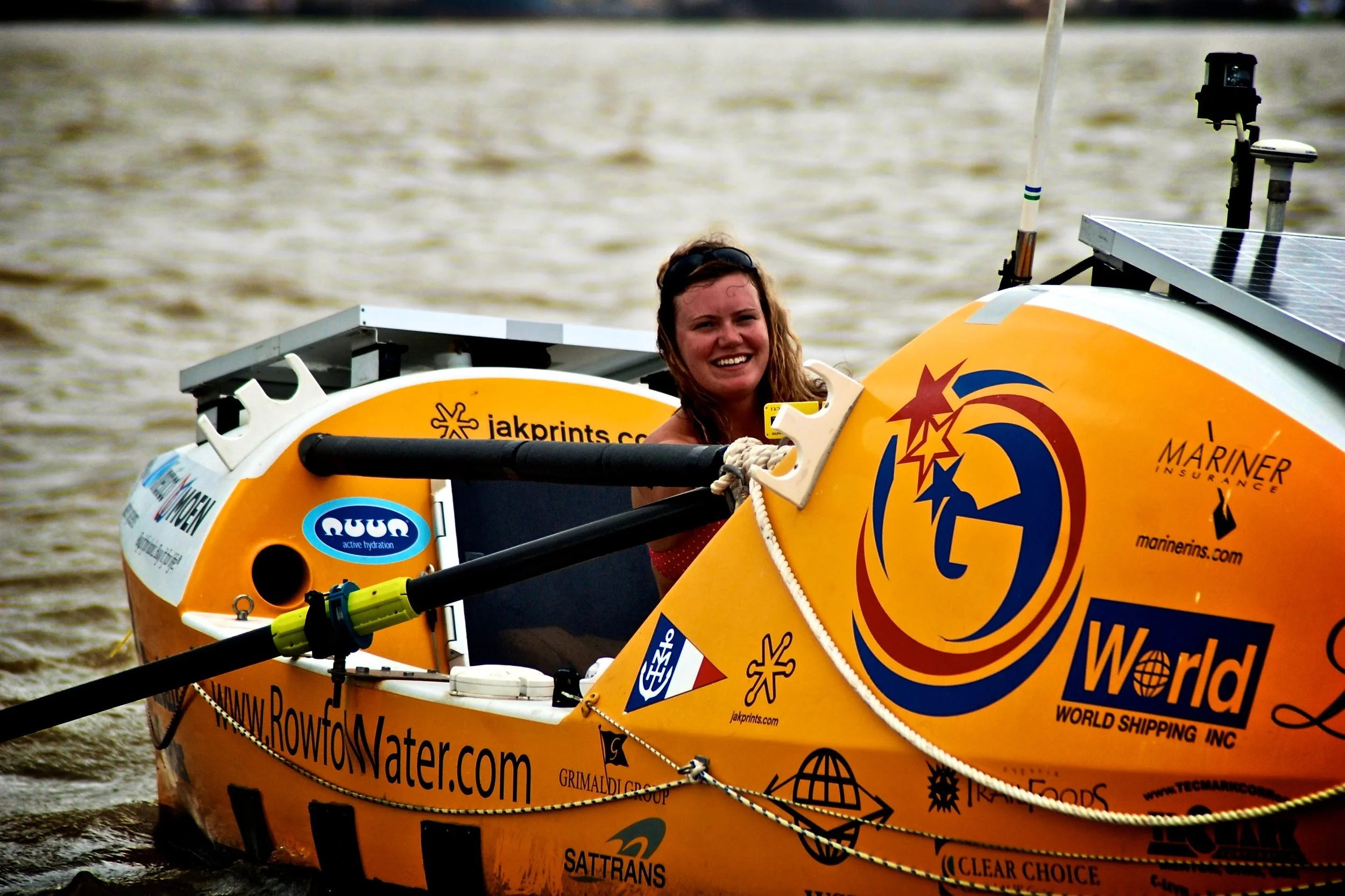 A smiling woman with wet hair in a yellow watercraft adorned with racing logos and sponsors, on a body of water.