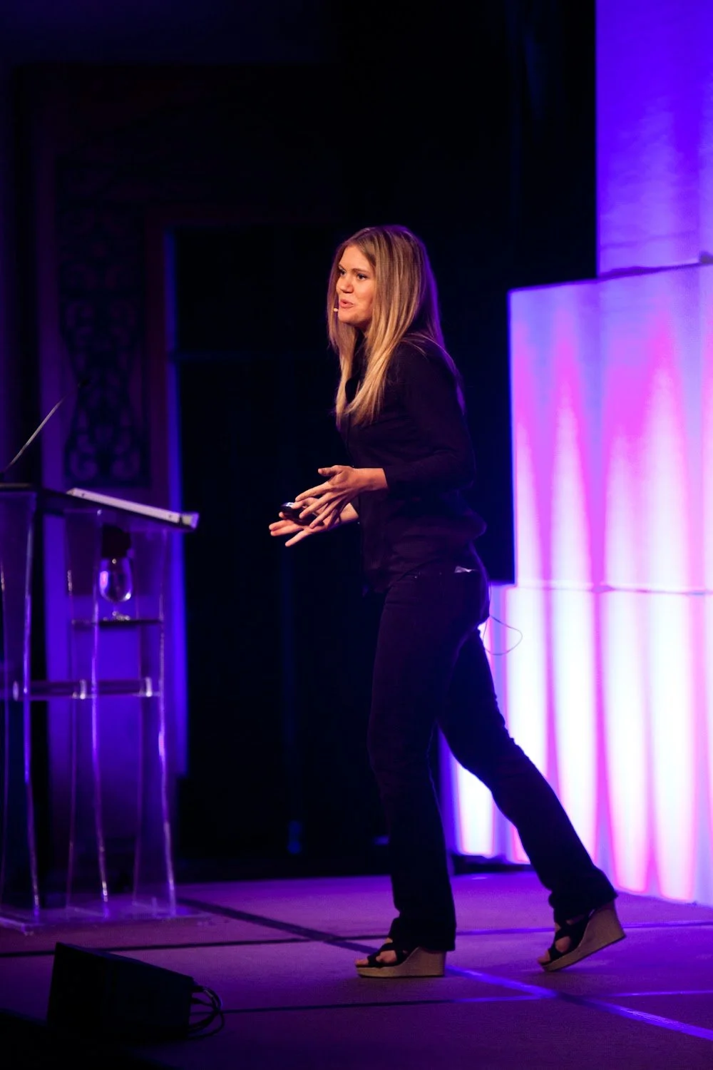 A woman with long blonde hair giving a presentation on stage with purple and pink lighting, standing next to a transparent lectern.