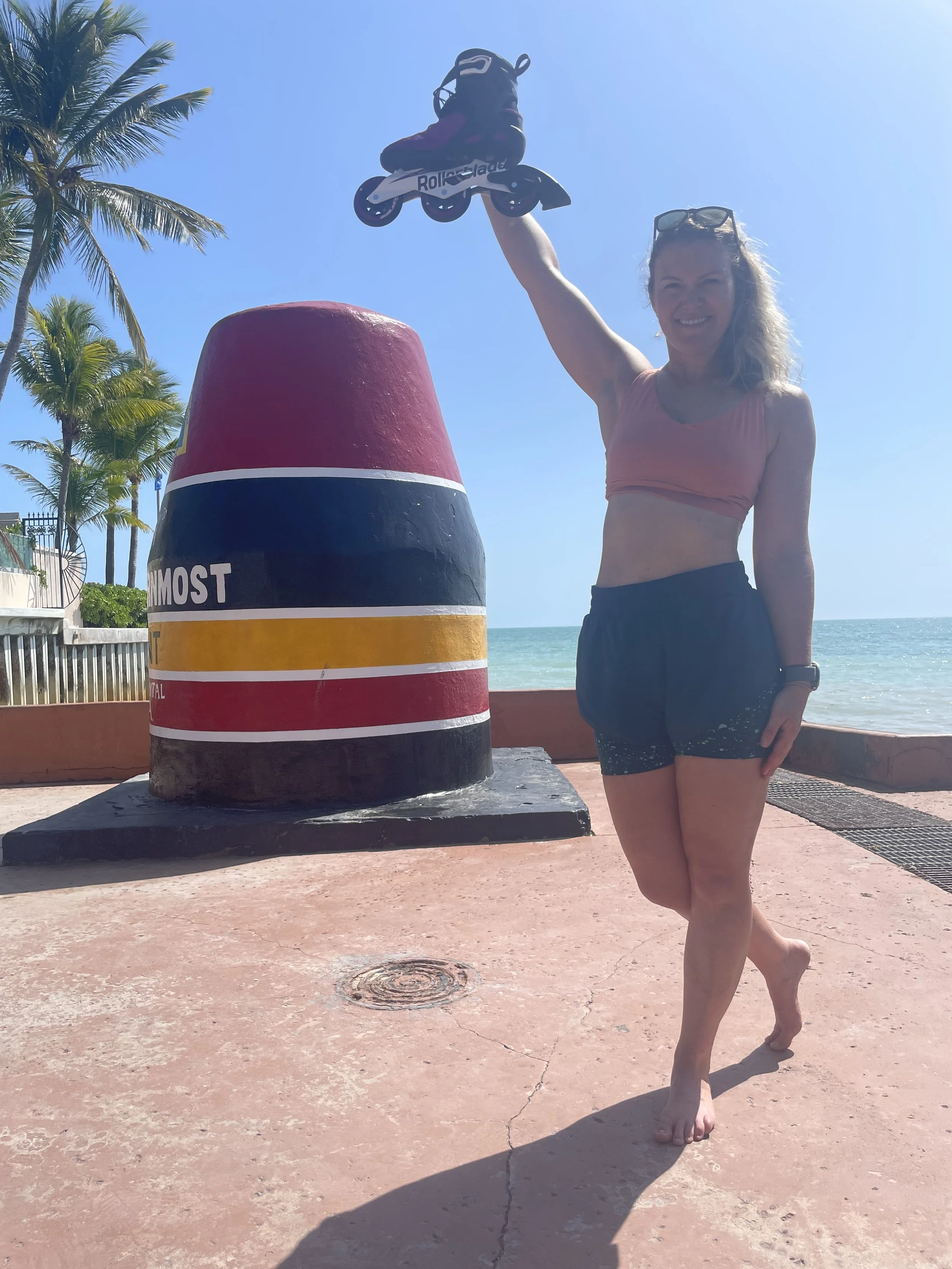 A smiling woman holding a rollerblade toy above her head at the beach, standing next to a large painted buoy with the ocean and palm trees in the background.