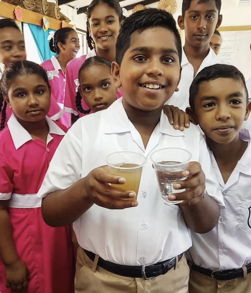 Group of school children smiling, wearing school uniforms, holding glasses of water and juice, in a classroom setting.