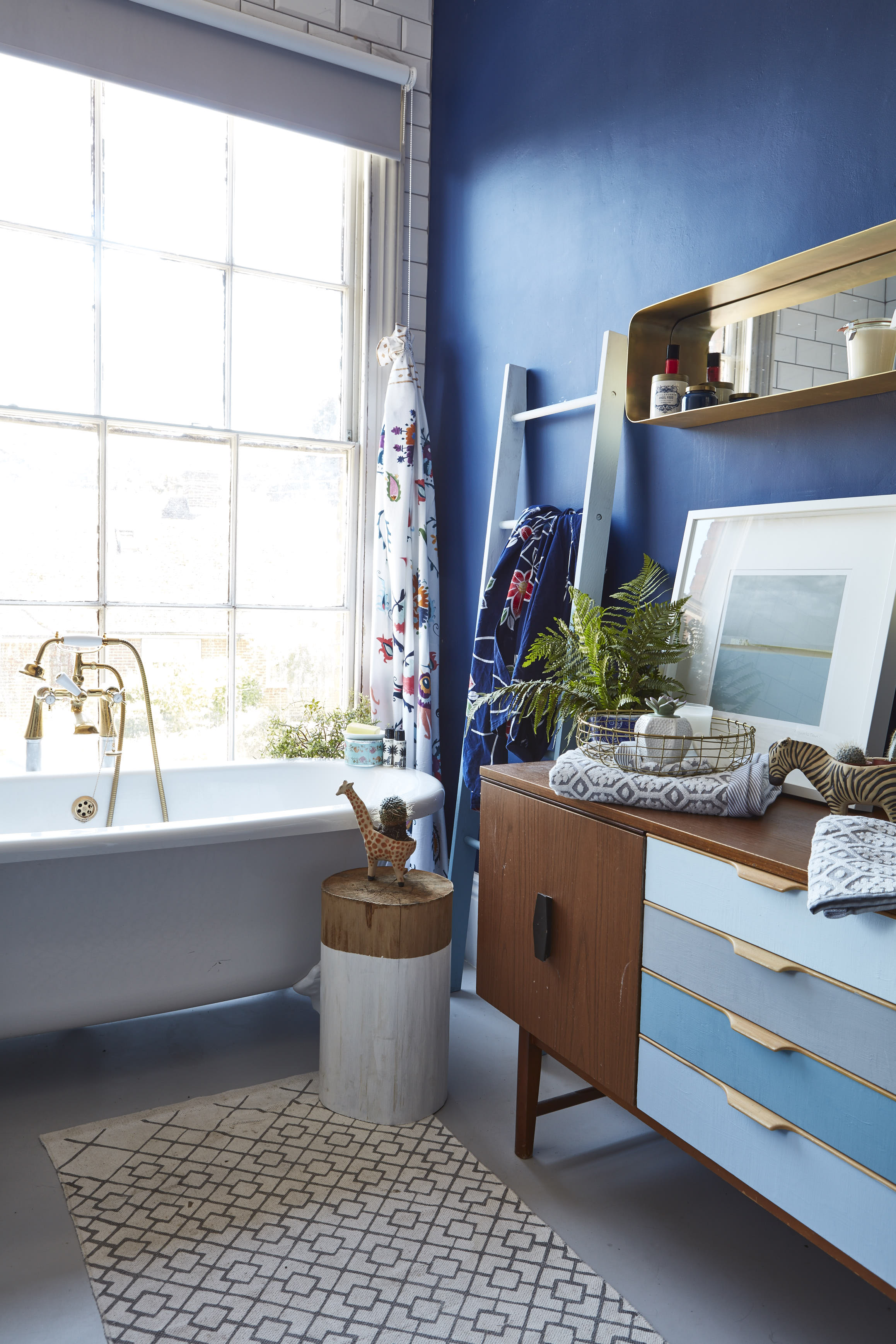 Traditional roll-top bath with gold fittings, Waverly range at Homebase . Sideboard, bought at a junk shop and painted in colours from the new Bert and May blue ranges. Rug, H+M. Towel ladder from Next, repainted in Bert + May colours. Paintings by artists from Dublin, Deirdre Hayden and Suffolk, Abi Fawcus . Selection of accessories painted in Strong White by Farrow and Ball. The wall is painted in Tuxedo Blue by Valspar at B&Q.