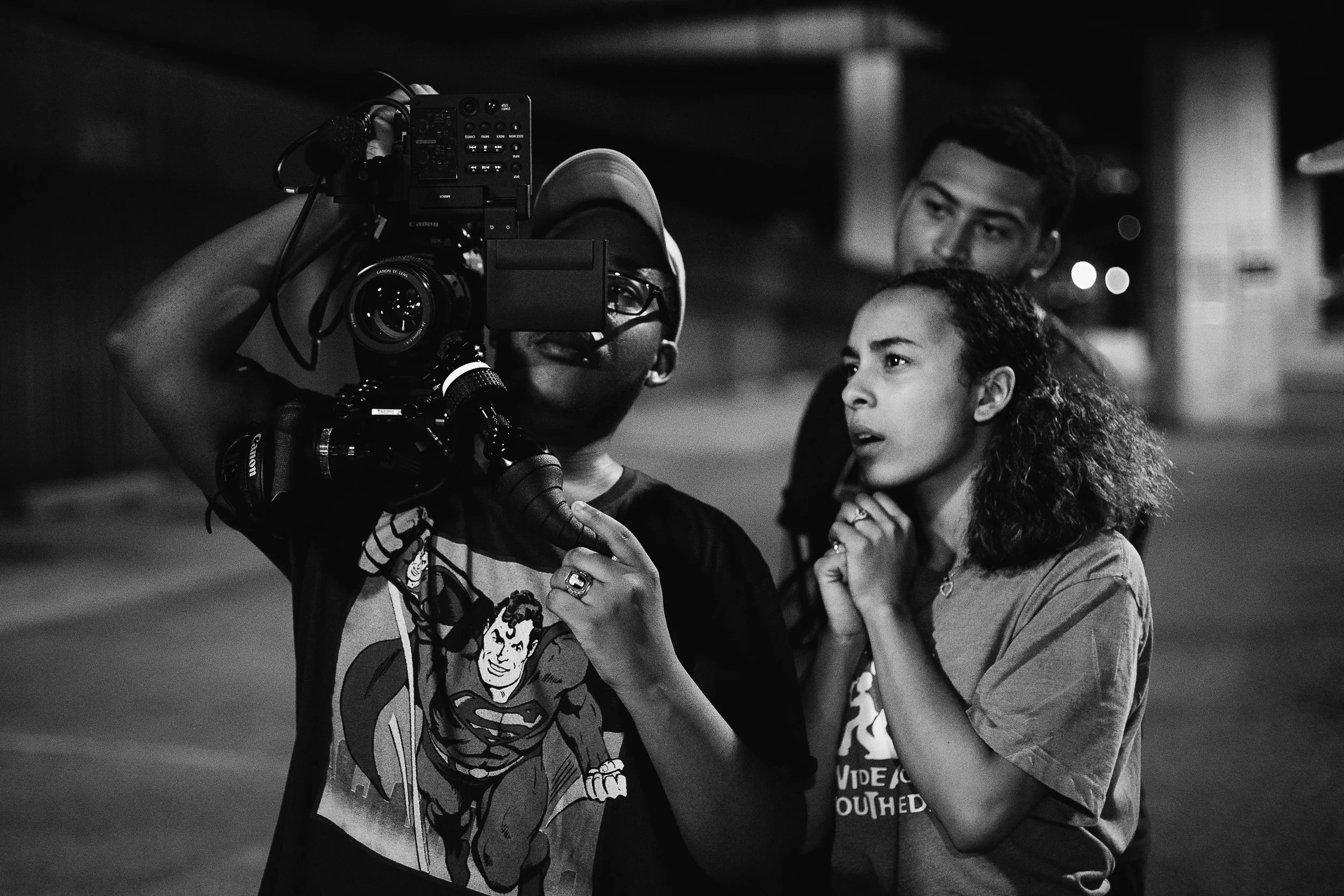 Three Black students are working with a big video camera outside at night.
