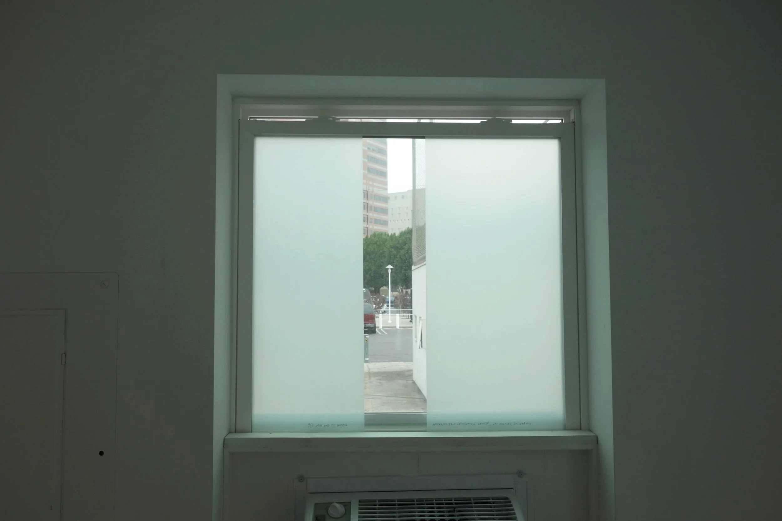 Installation view: Ashley Hunt, 955 Men and 55 Women, Metropolitan Detention Center, Los Angeles , 2014. Acetate on glass framing a view of the nearby Metropolitan Detention Center.
