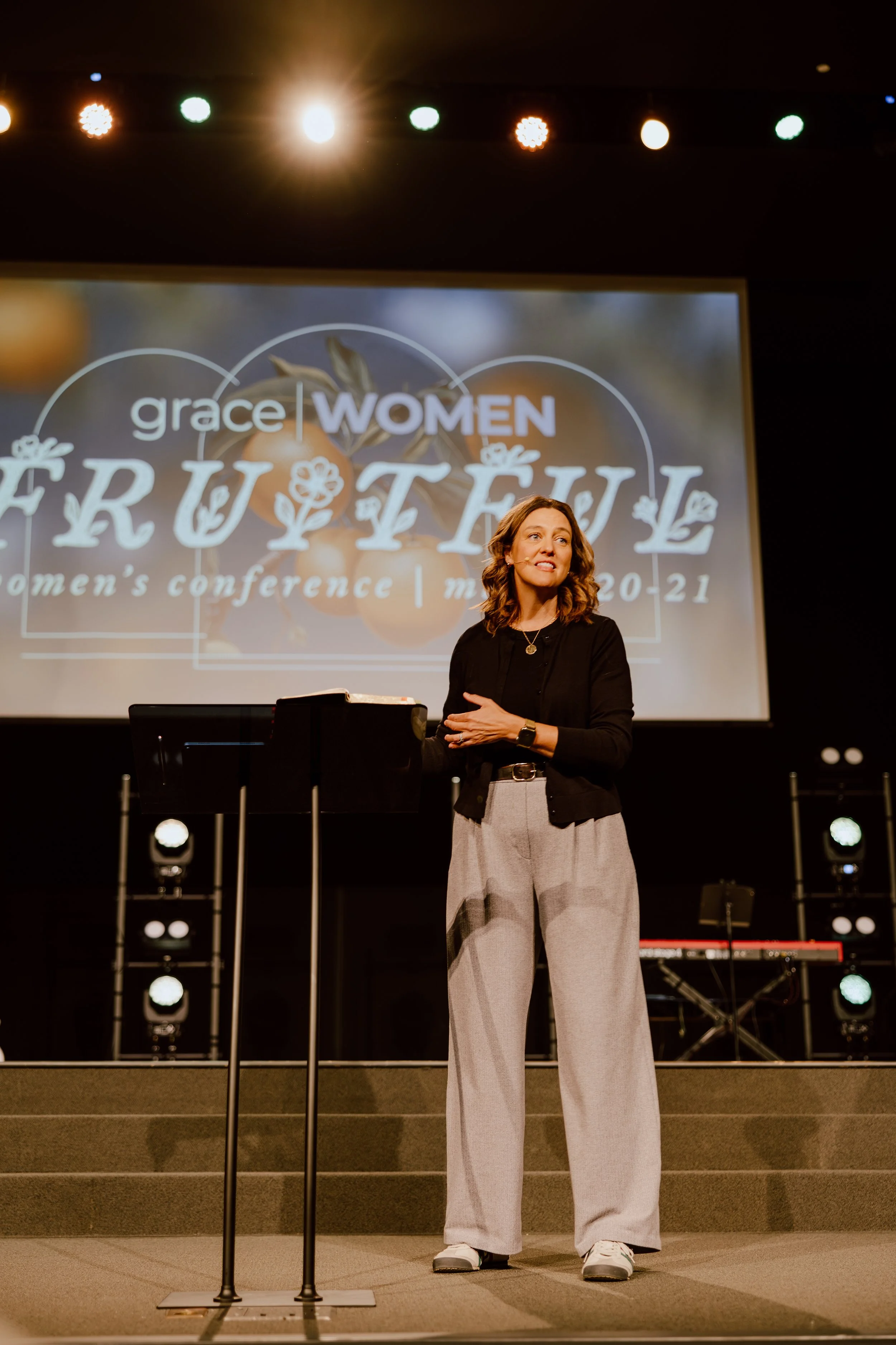 A woman standing on stage in front of a large screen, giving a presentation at a conference titled 'Grace Women'. She is wearing a black top, beige wide-leg pants, and sneakers, with her hair styled in loose waves.