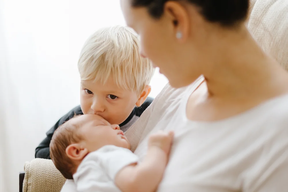 big brother kissing baby brother while mom holds baby in the rocking chair