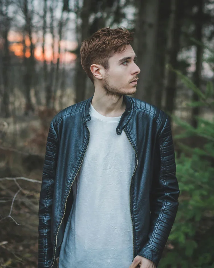 A young man with light brown hair and facial hair wearing a black leather jacket and white T-shirt, standing in a forest during sunset, looking to the right.