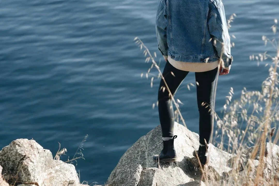 Person standing on rocks by a body of water, gazing at it, wearing a denim jacket, leggings, and boots.